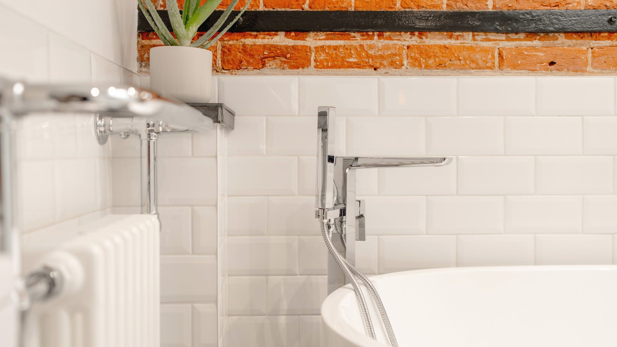 A modern bathroom corner featuring a white bathtub with a silver faucet, adjacent to a white radiator. the wall combines clean white tiles and exposed brick, topped with a small potted plant.