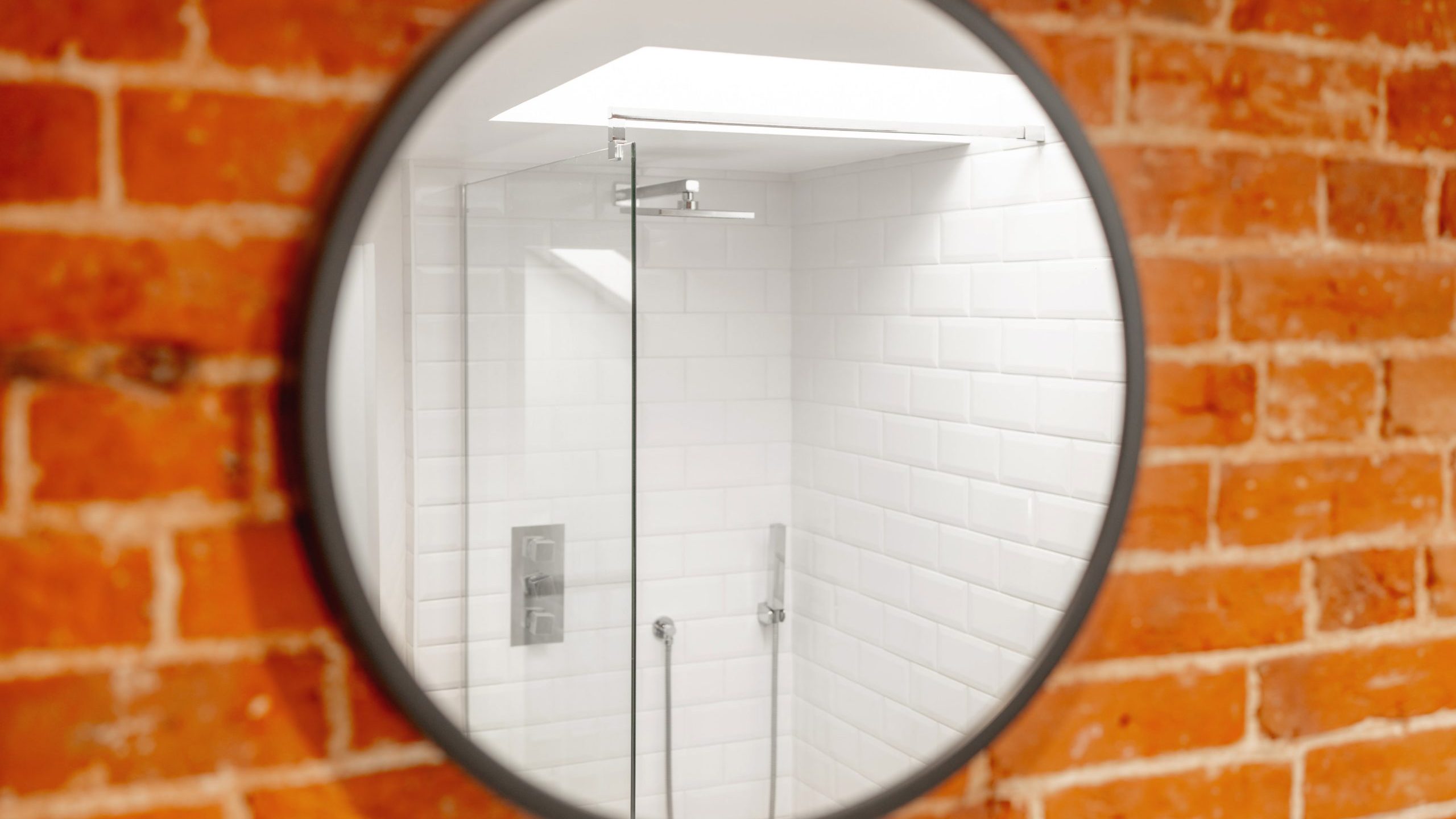 A circular mirror reflects an image of a white-tiled shower area, mounted against a rustic brick wall. the shower features a glass door and stainless steel fixtures.
