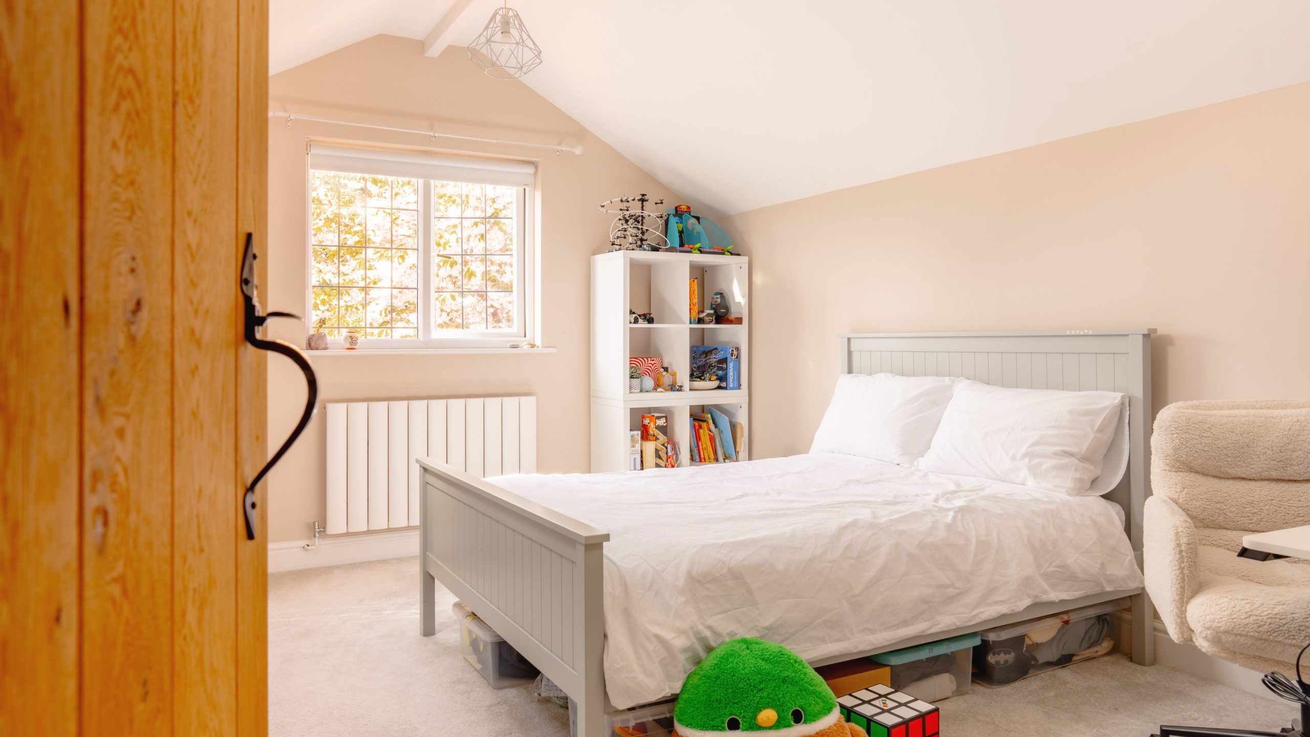A cozy attic bedroom features a double bed with white bedding, a window with natural light, shelving with toys, and a green plush toy at the foot of the bed.