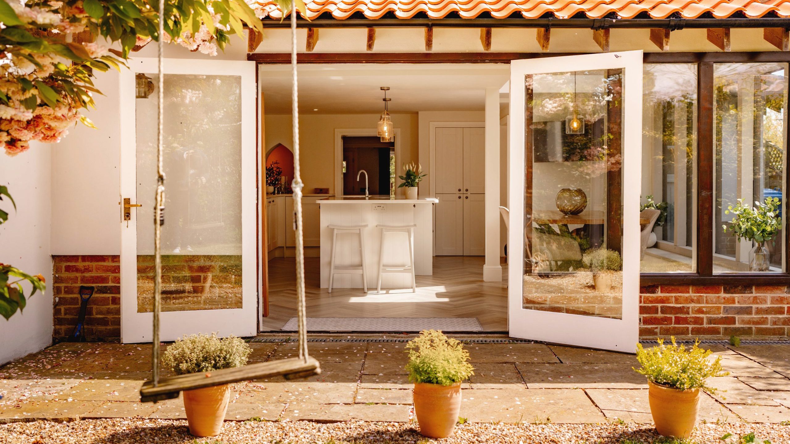 A bright and sunny view of a home's interior visible through large open glass doors, showcasing a kitchen island and modern decor, framed by lush outdoor greenery and potted plants.
