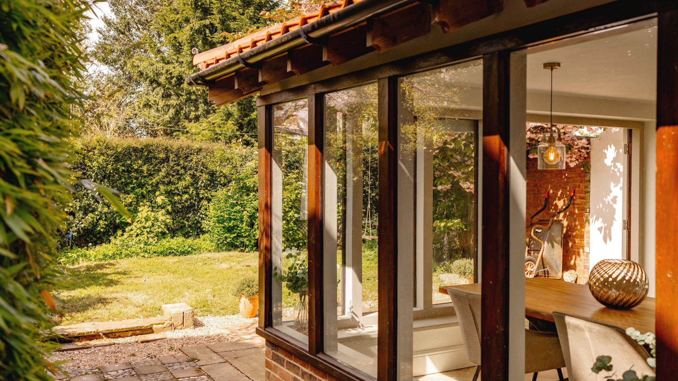 View of a sunlit garden through open glass doors of a cozy home with visible dining area inside, featuring warm, inviting decor and surrounded by lush greenery.