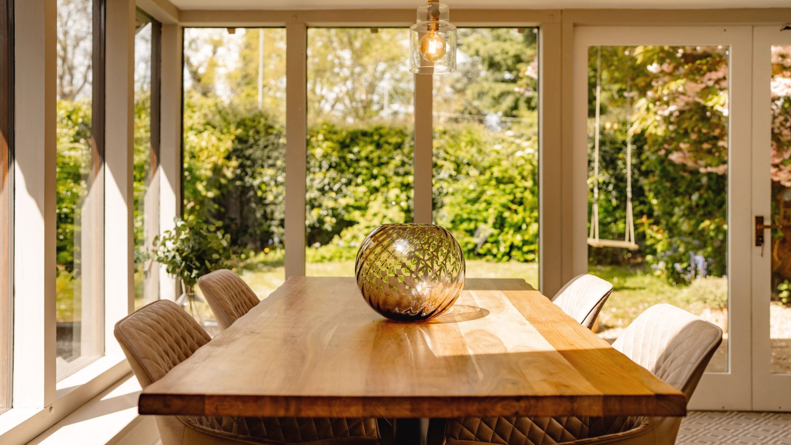 A bright dining room with a wooden table and tufted chairs. a decorative glass vase sits in the center. large windows offer a view of a garden, and a hanging light fixture adds a cozy ambiance.
