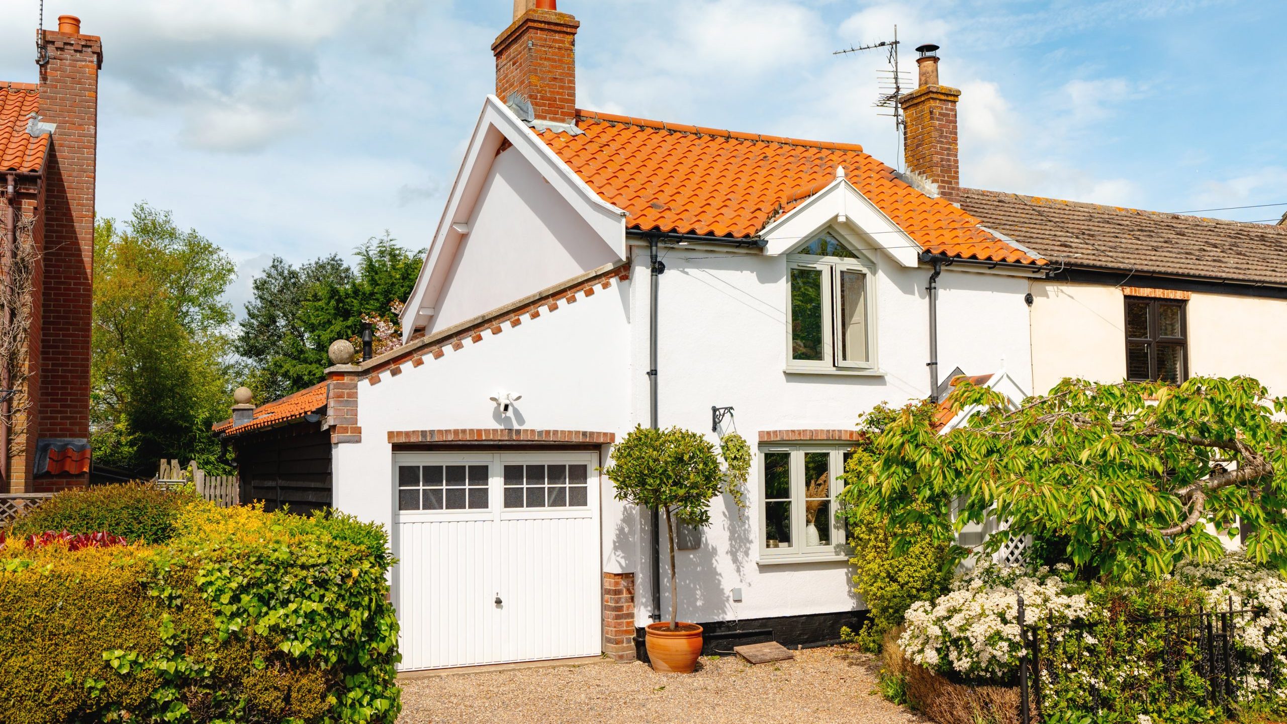 A quaint, well-kept house with a white facade and orange tiled roof under a clear blue sky, featuring a garage, a chimney, and lush greenery around.