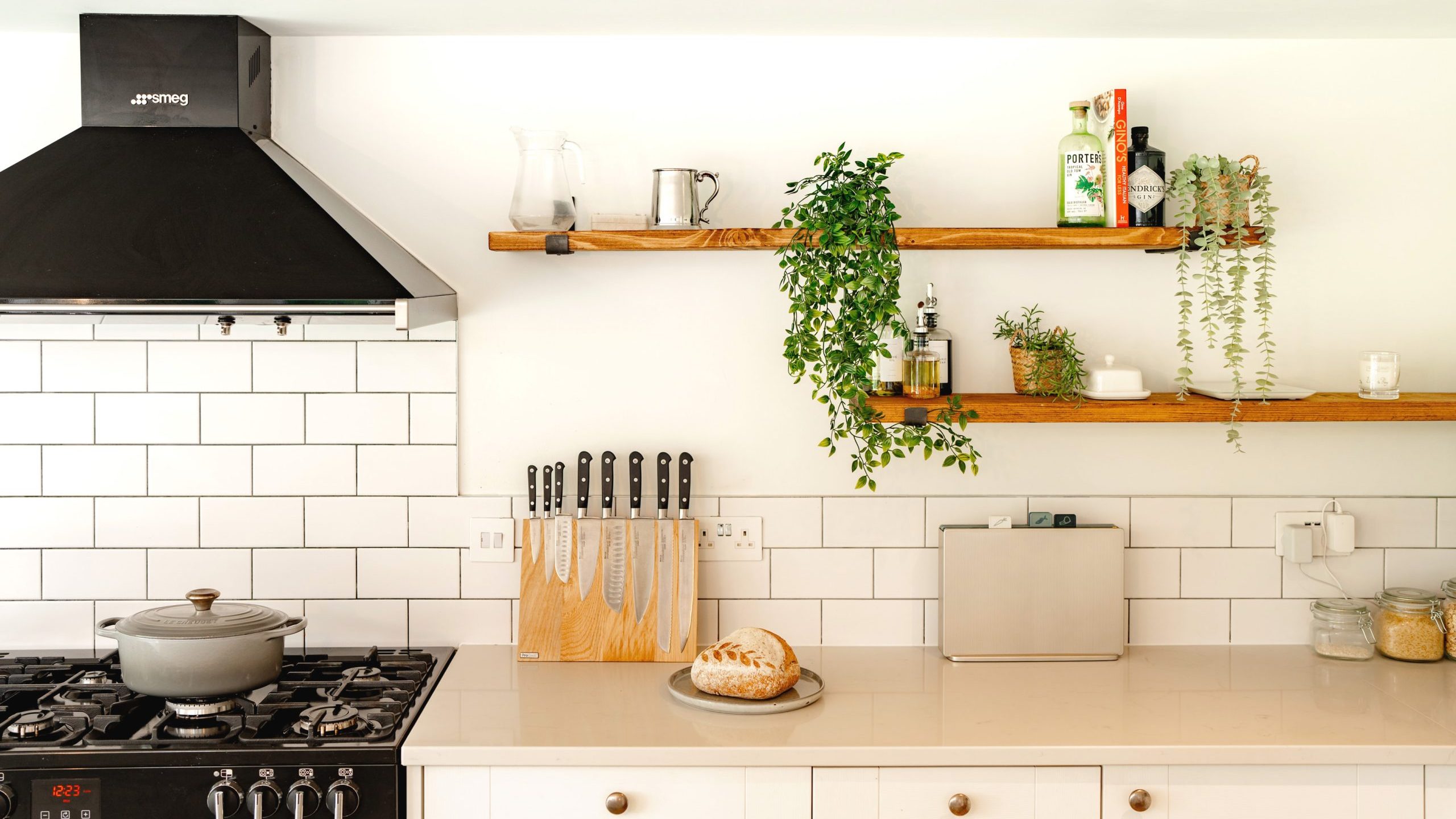 A modern kitchen with white cabinetry and black countertop, featuring a gas stove, a wall-mounted wooden shelf with plants and spices, and hanging utensils.