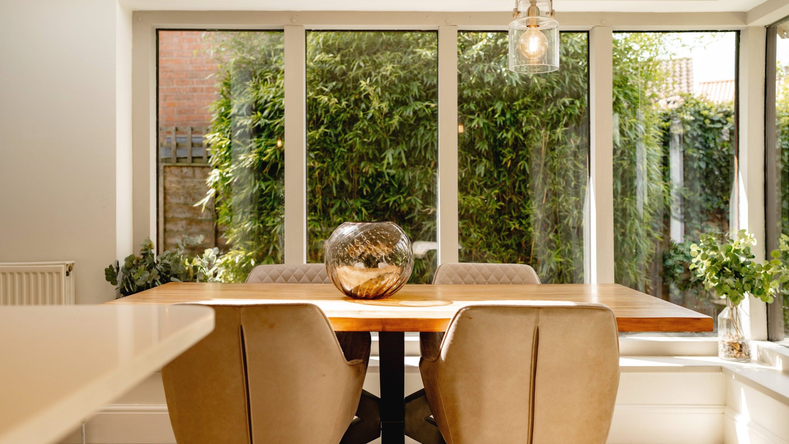A modern dining room with a wooden table, tan chairs, and a large window overlooking a lush garden. natural light fills the room, highlighting a decorative glass bowl on the table.