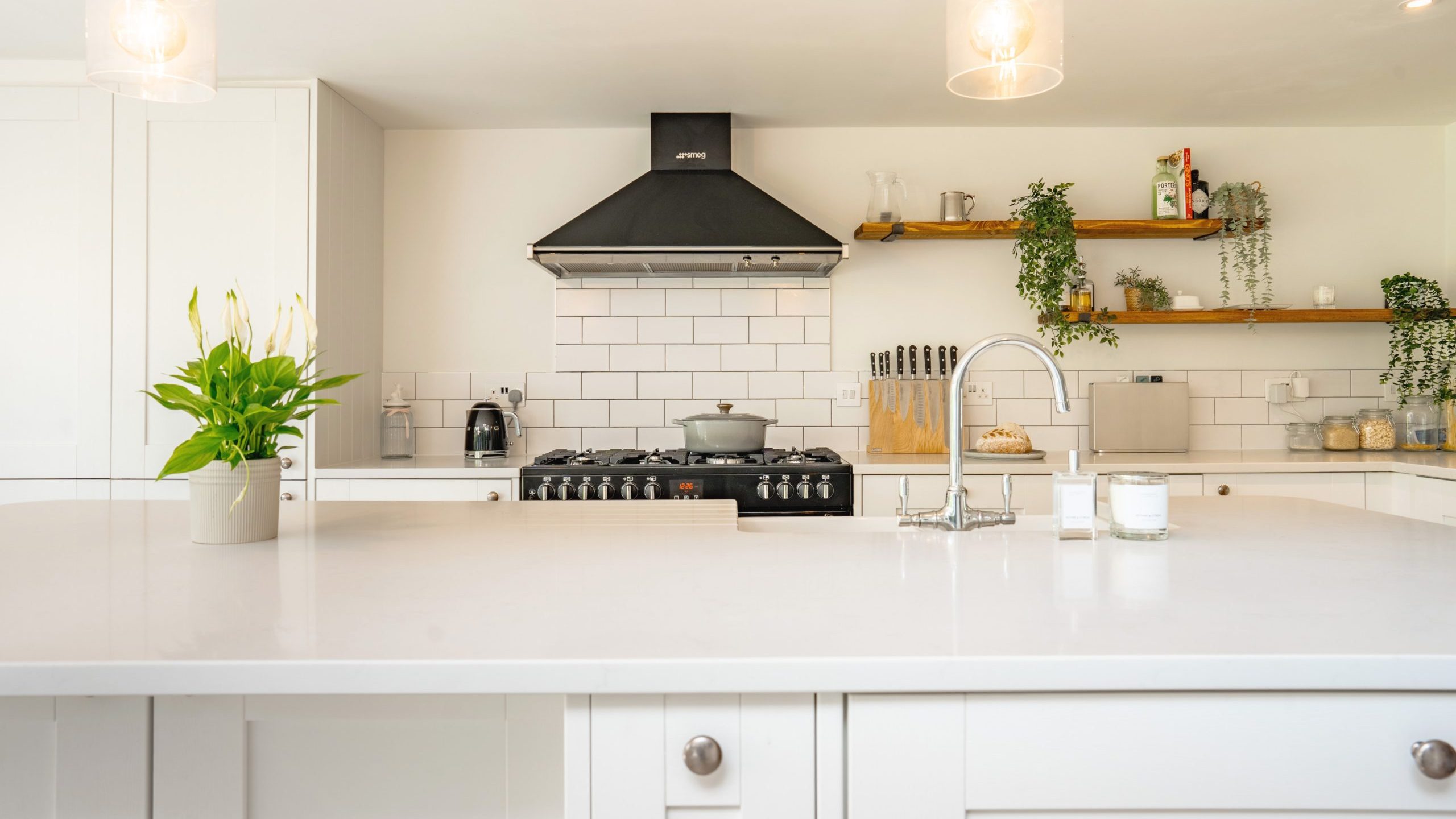 Modern kitchen interior with white cabinetry, a central island with a gas stove, hanging pendant lights, and green plants on the shelves. the room is well-lit and features a clean, minimalistic design.