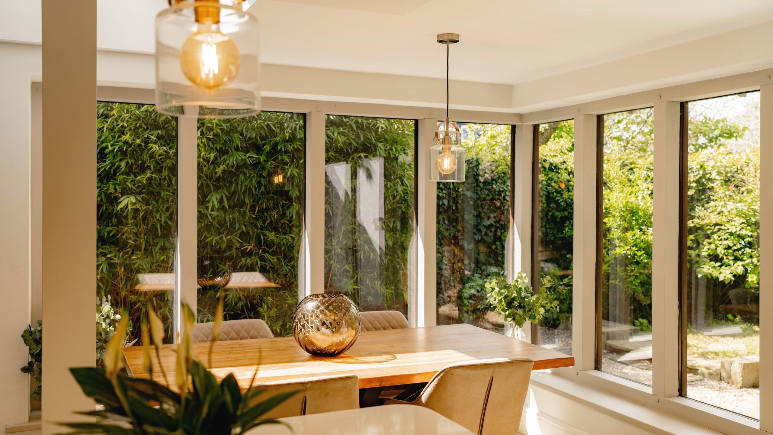 A bright, modern dining room with large windows overlooking a garden. the room features a wooden table, chairs, and stylish hanging lights.