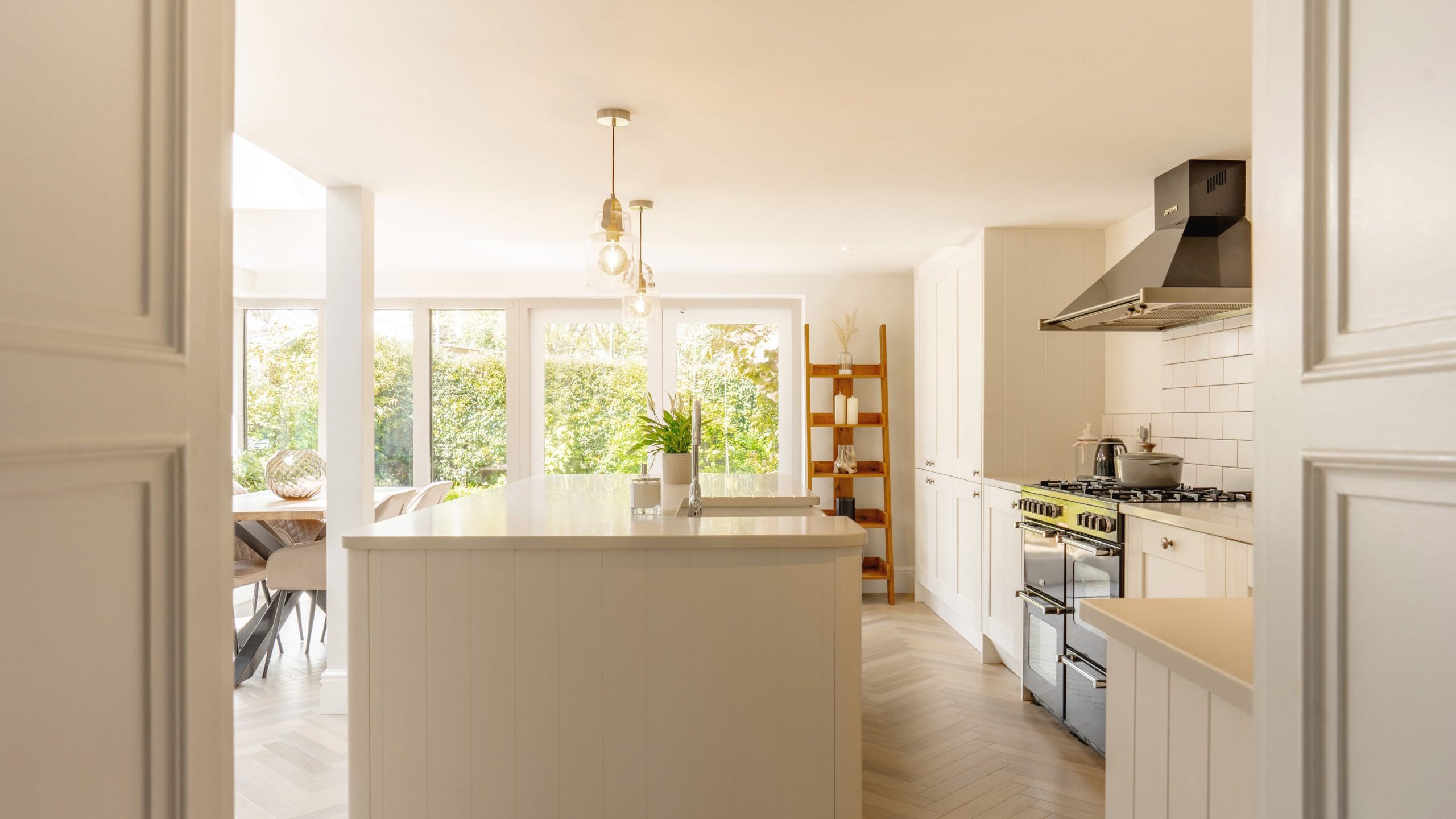 A bright, modern kitchen with white cabinetry and stainless steel appliances. the room has hardwood floors and opens up to a dining area with large windows overlooking a garden.