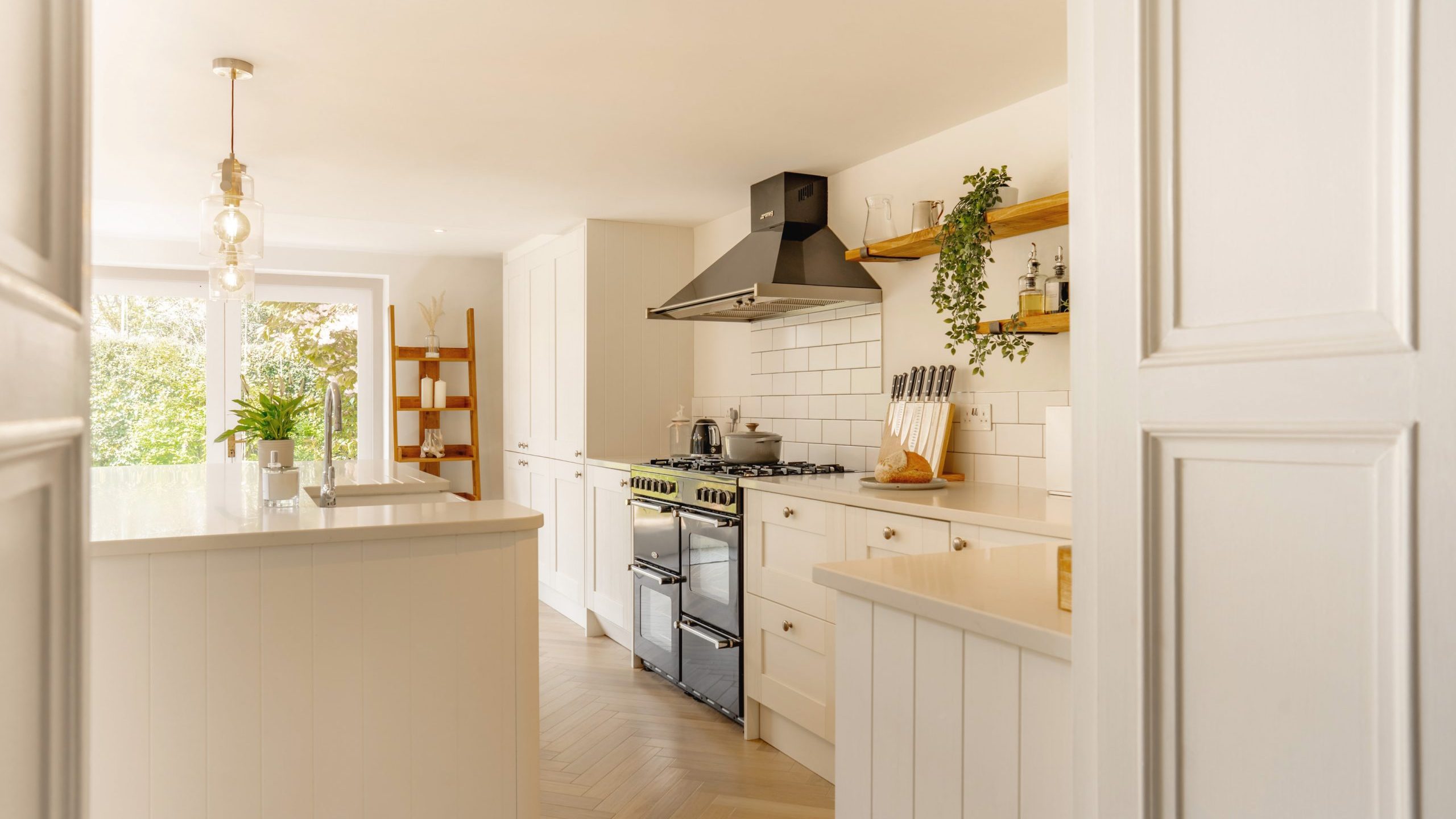 A modern, bright kitchen with white cabinetry and stainless steel appliances, featuring a gas stove, a hood, and a wooden floor, viewed from a doorway.