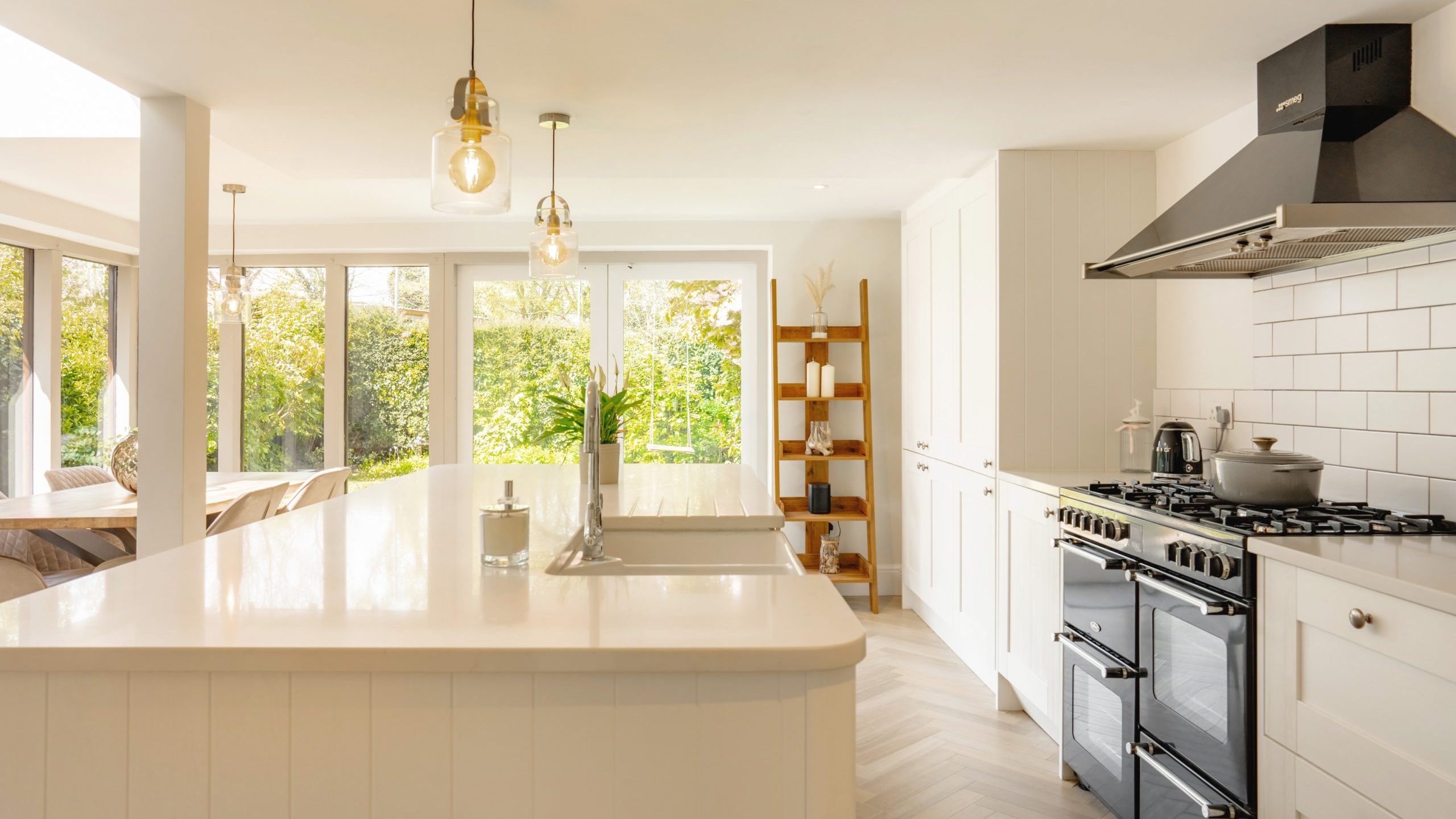 A bright, modern kitchen with white cabinetry and subway tiles, featuring stainless steel appliances, a kitchen island, pendant lights, and a view of a garden through large windows.