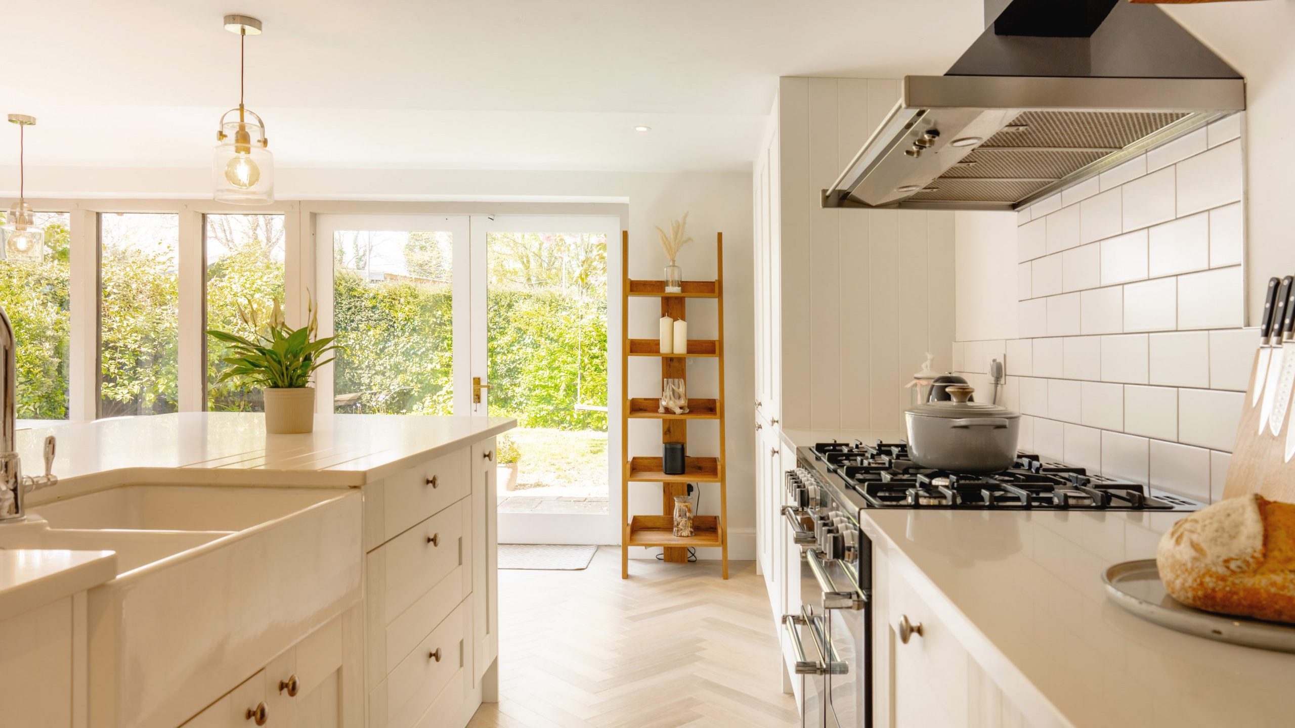 A modern kitchen with white cabinetry and stainless steel appliances. the room is bright with natural light from large windows overlooking a green garden. a gas stove, a cooking pot, and green plants are visible.