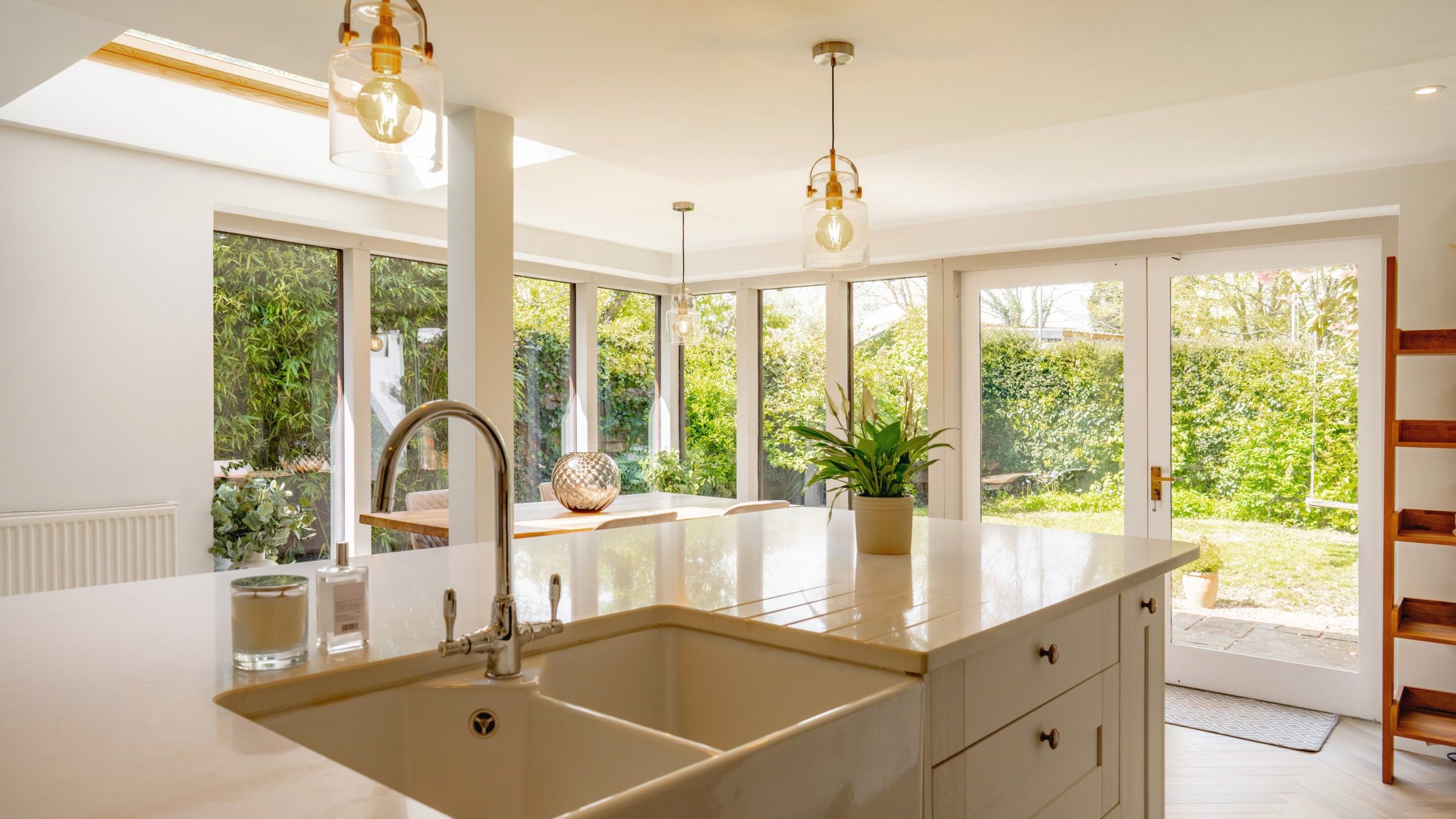 A spacious, well-lit modern kitchen featuring a large island with a white farmhouse sink, pendant lights, and a view of the garden through glass doors.