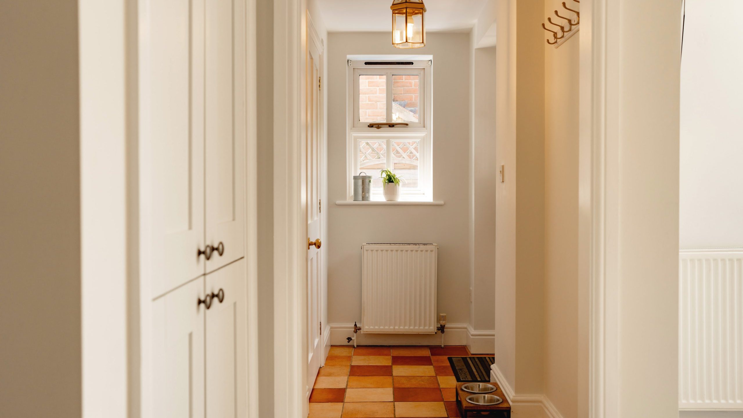 Narrow hallway in a home, featuring terracotta tiled flooring, white walls, and a hanging lantern light. a window at the end provides natural light, with a radiator and shelves on the side.