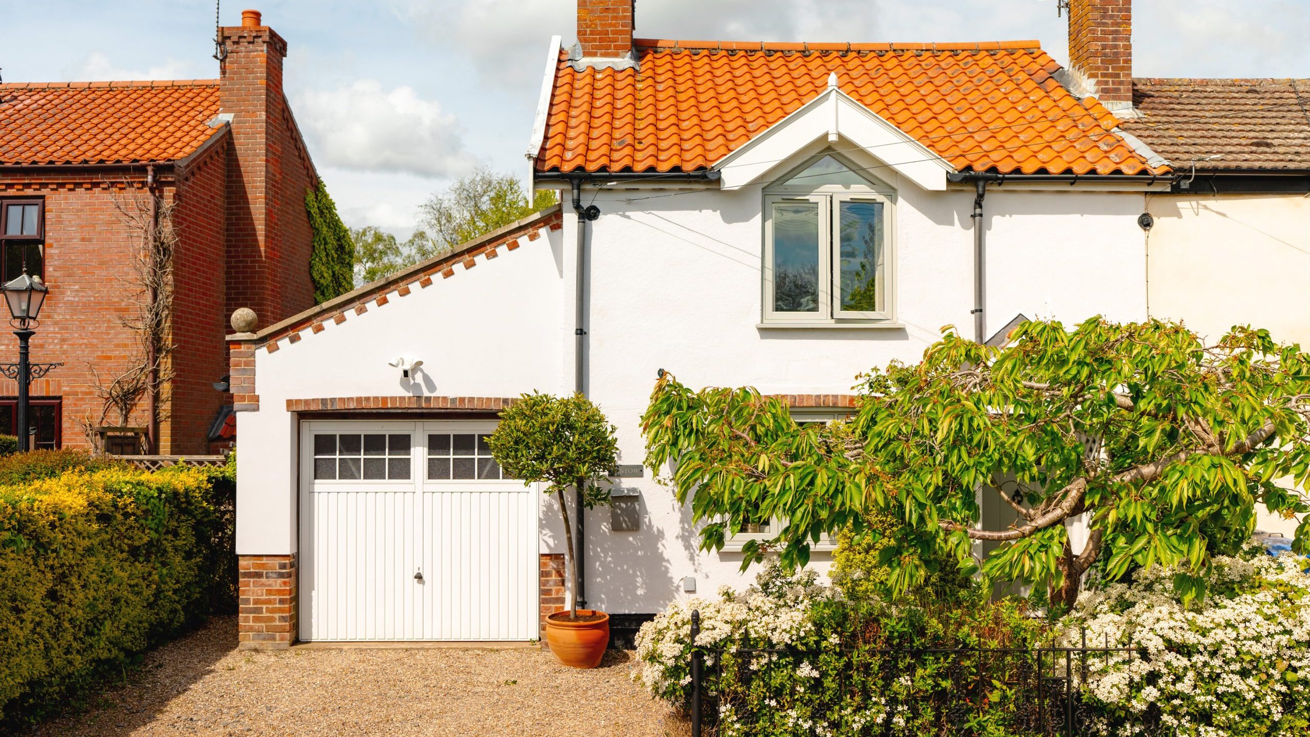 A quaint home with a white garage door and a terra cotta pot by the driveway. lush greenery surrounds the house, and a cat sits on the roof under a sunny sky.