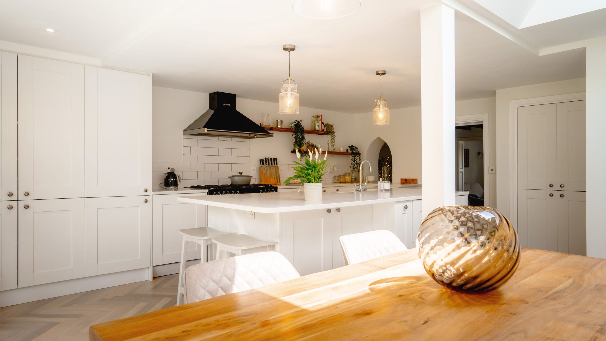 A modern kitchen with white cabinetry, a central island with bar stools, and pendant lights. a wooden dining table is in the foreground and the space is well-lit with natural light.