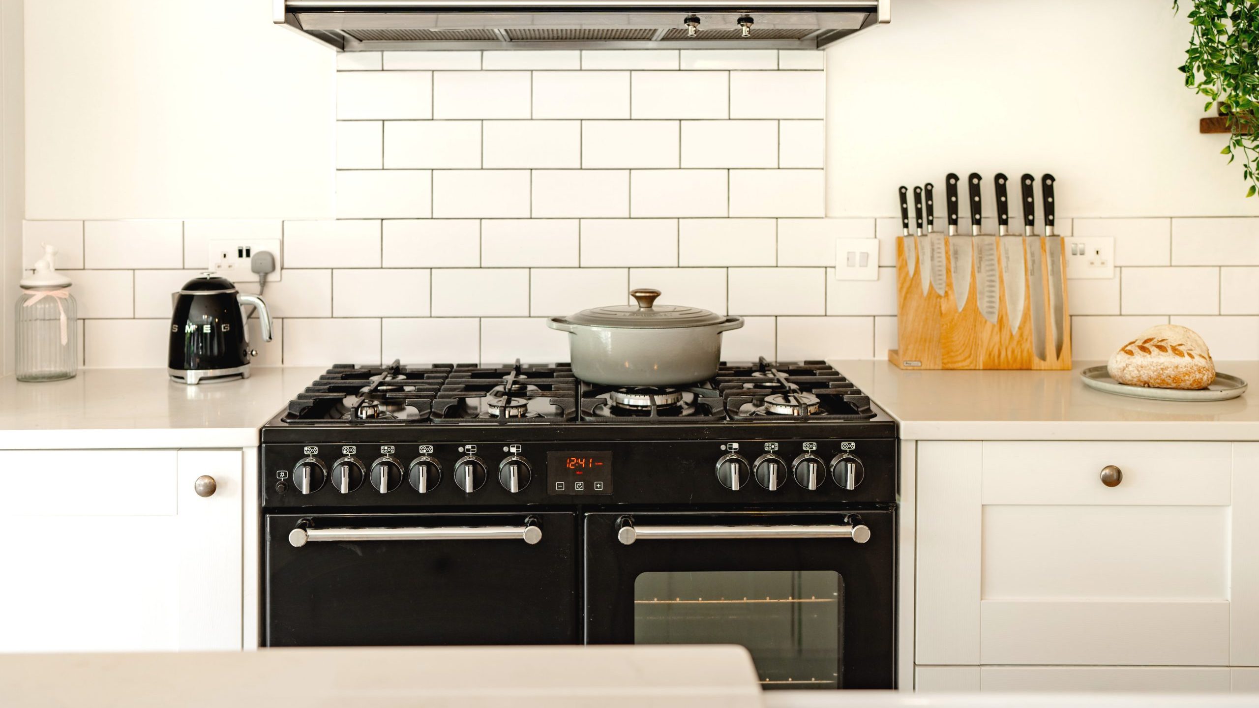 A modern kitchen featuring a clean, white-tile backsplash, and a stainless steel stove with a gray pot on top. there's a bread loaf and knife set on the counter, and wooden shelves above the stove.