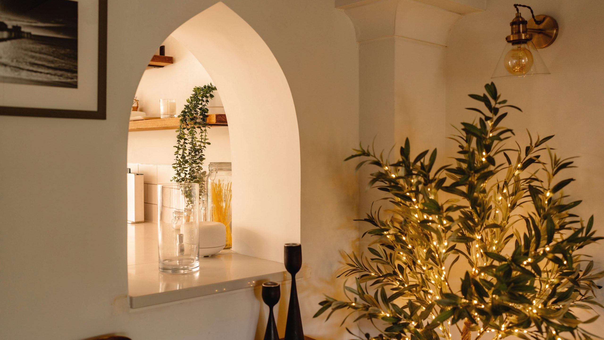 Interior of a cozy room featuring warm lighting, an arched doorway, and a view into an adjoining space with a shelf and decorative items, including plants and a small tree illuminated by string lights.