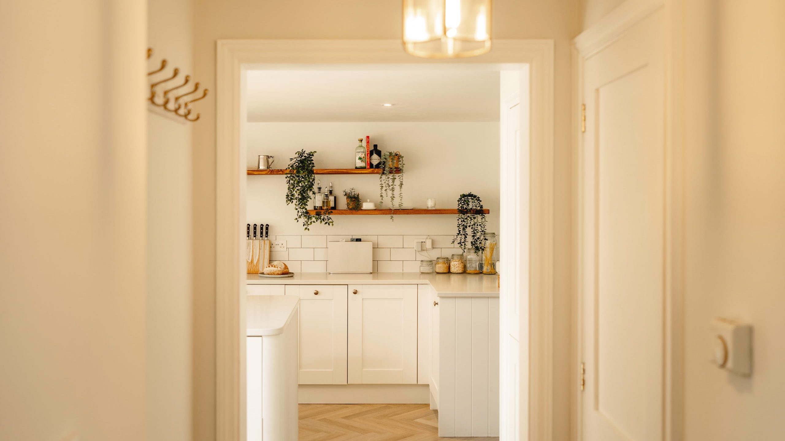 A bright, modern kitchen viewed through a doorway, featuring white cabinetry, wooden countertops, and decorative plants, illuminated by natural light and a hanging light fixture.
