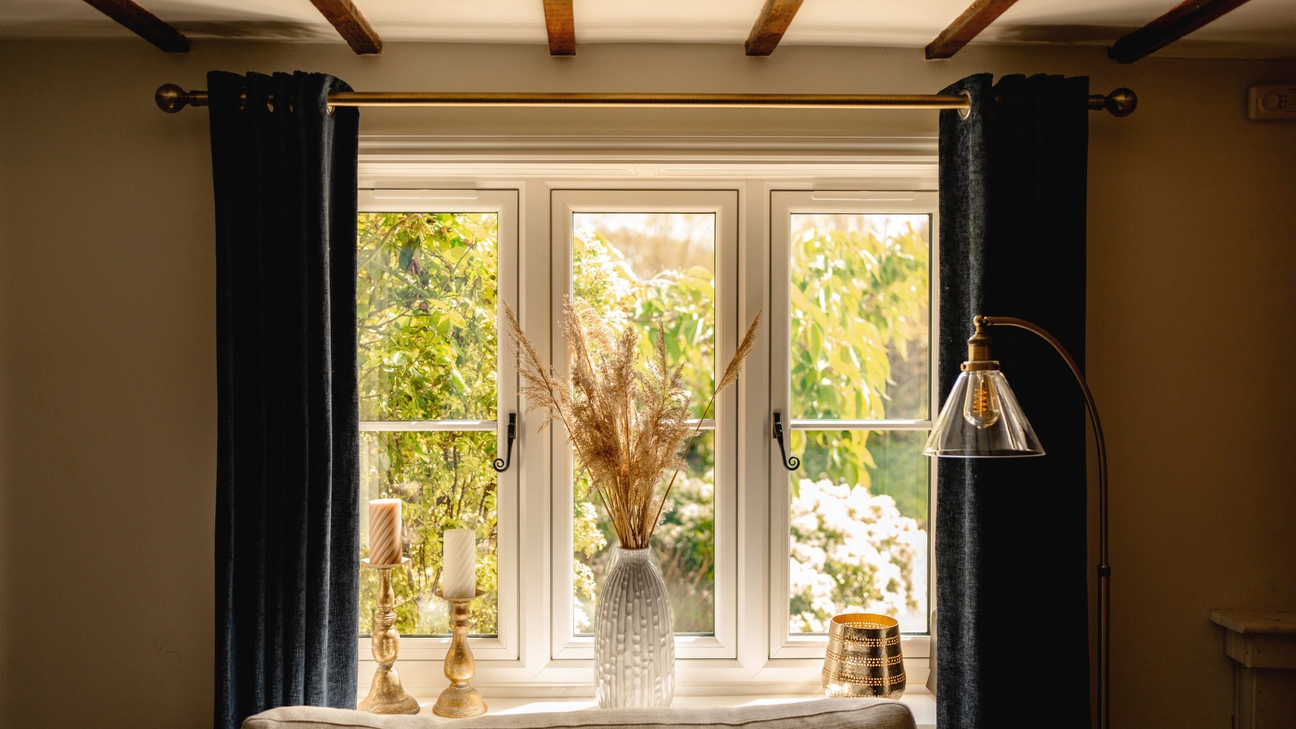 A cozy room interior with sunlight streaming through a window framed by dark curtains. a decorative table holds a vase with dried plants, illuminated by a lamp, with a lush garden visible outside.