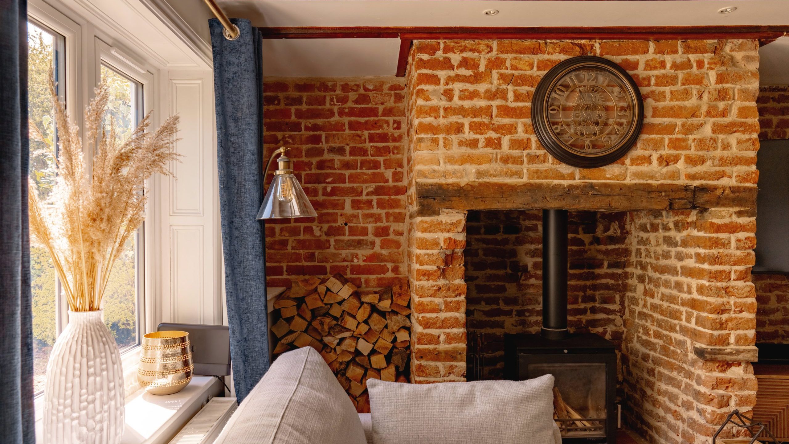 Cozy room featuring a brick fireplace with stacked wood, a plush sofa, and decorative items including a woven basket and dried plants beside a bright window.