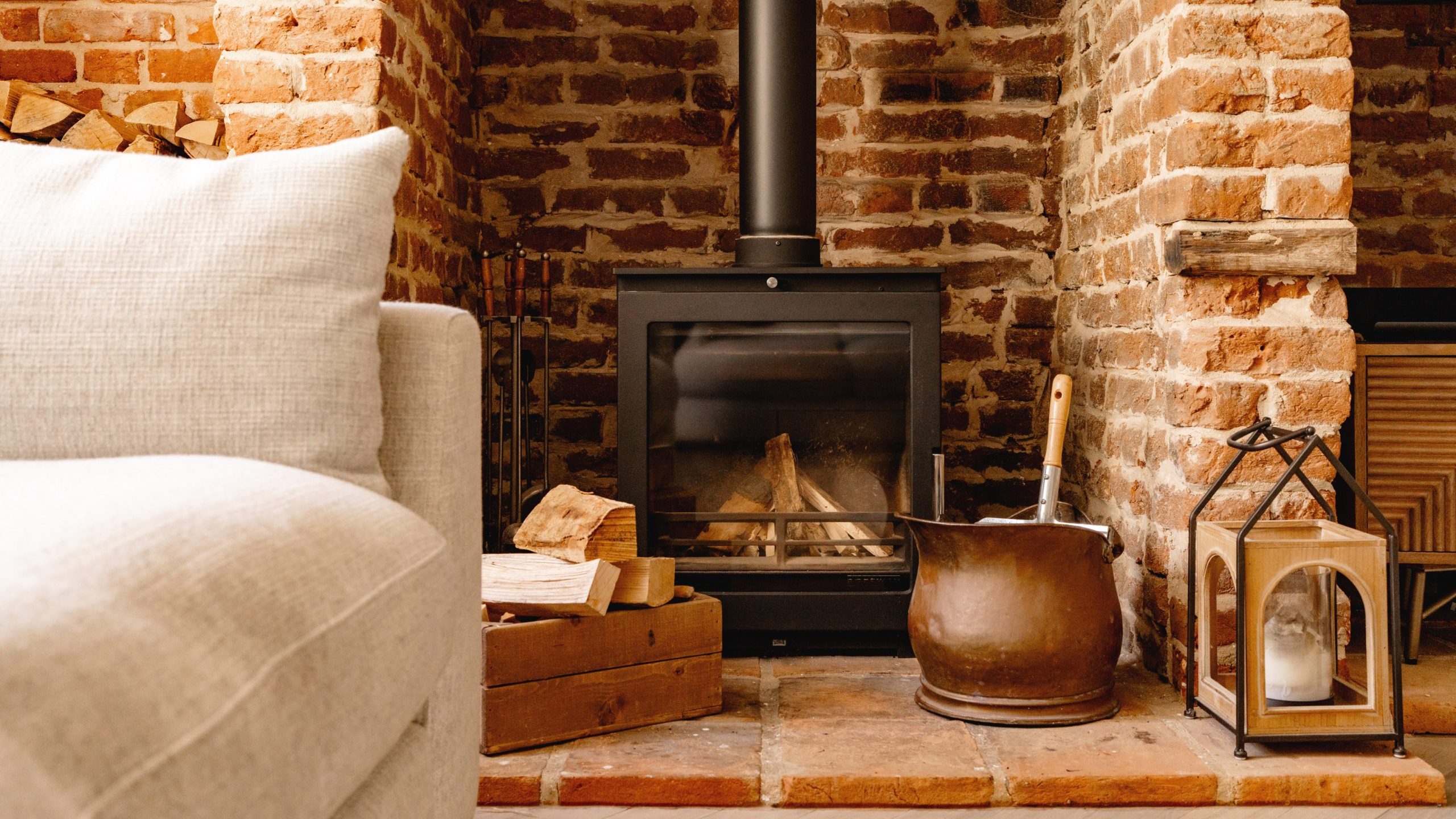 A cozy room corner featuring a wood-burning stove with visible flames, set against a red brick wall. there is a plush sofa, a wooden box with firewood, a metal pot, and a small lantern nearby.