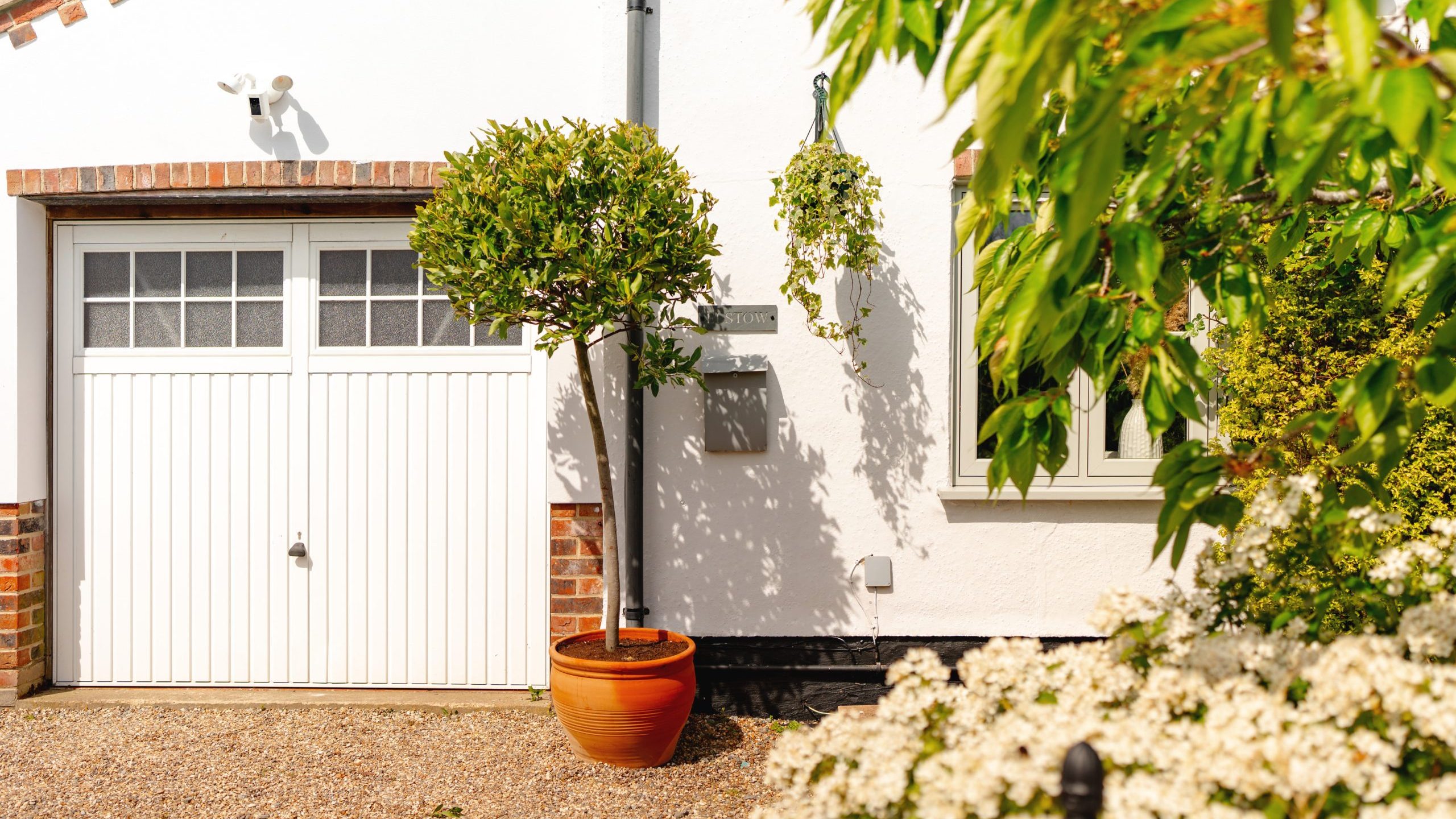 A bright and sunny view of a tidy white garage with double doors, flanked by a potted tree and brick details, next to a white residential home partly obscured by lush green foliage and white flowers in the foreground.