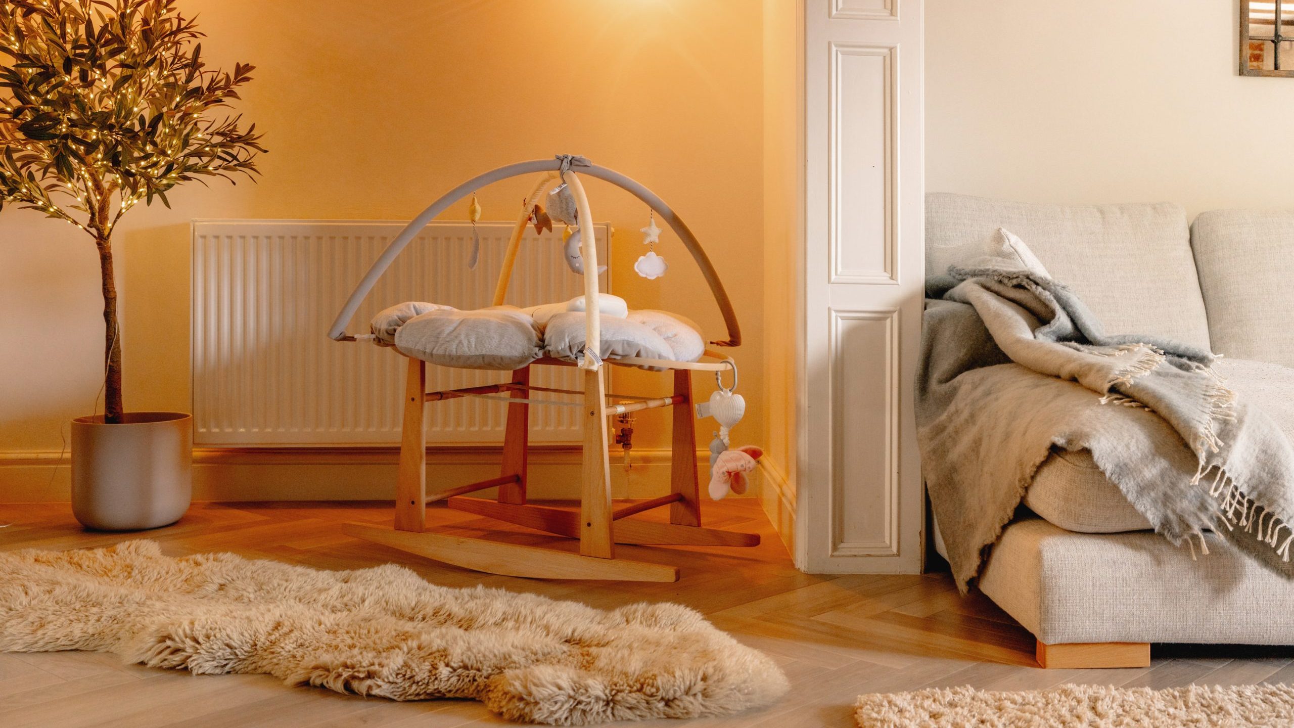 A cozy living room with a soft, glowing light featuring a wooden baby crib, a couch with a gray blanket, a potted tree, and a fluffy rug on wooden flooring.