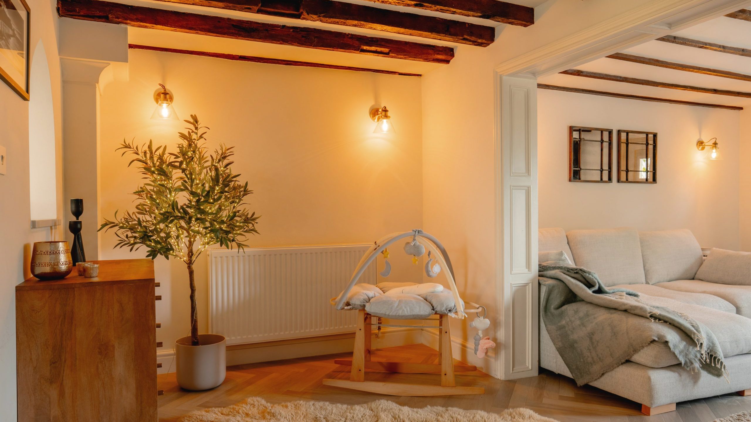 A cozy living room with warm lighting, featuring exposed wooden beams, a plush sofa covered with a gray blanket, a stylish rocking chair, and a potted indoor tree.