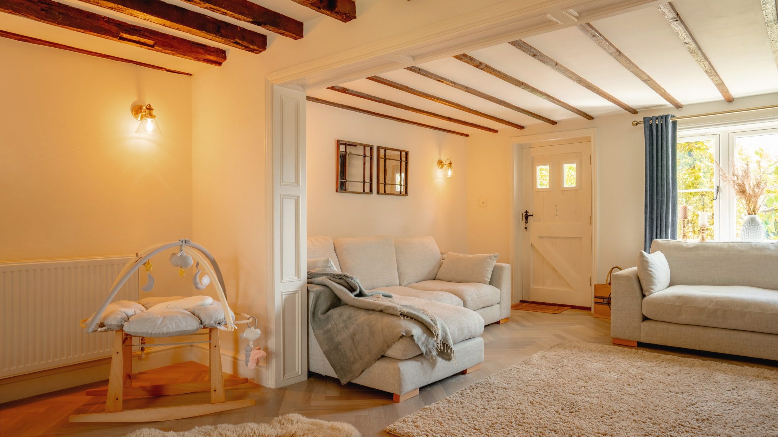 Warm, cozy living room featuring exposed wooden beams, two white sofas with a gray blanket, a large beige rug, and a wooden rocking horse near a bright window.