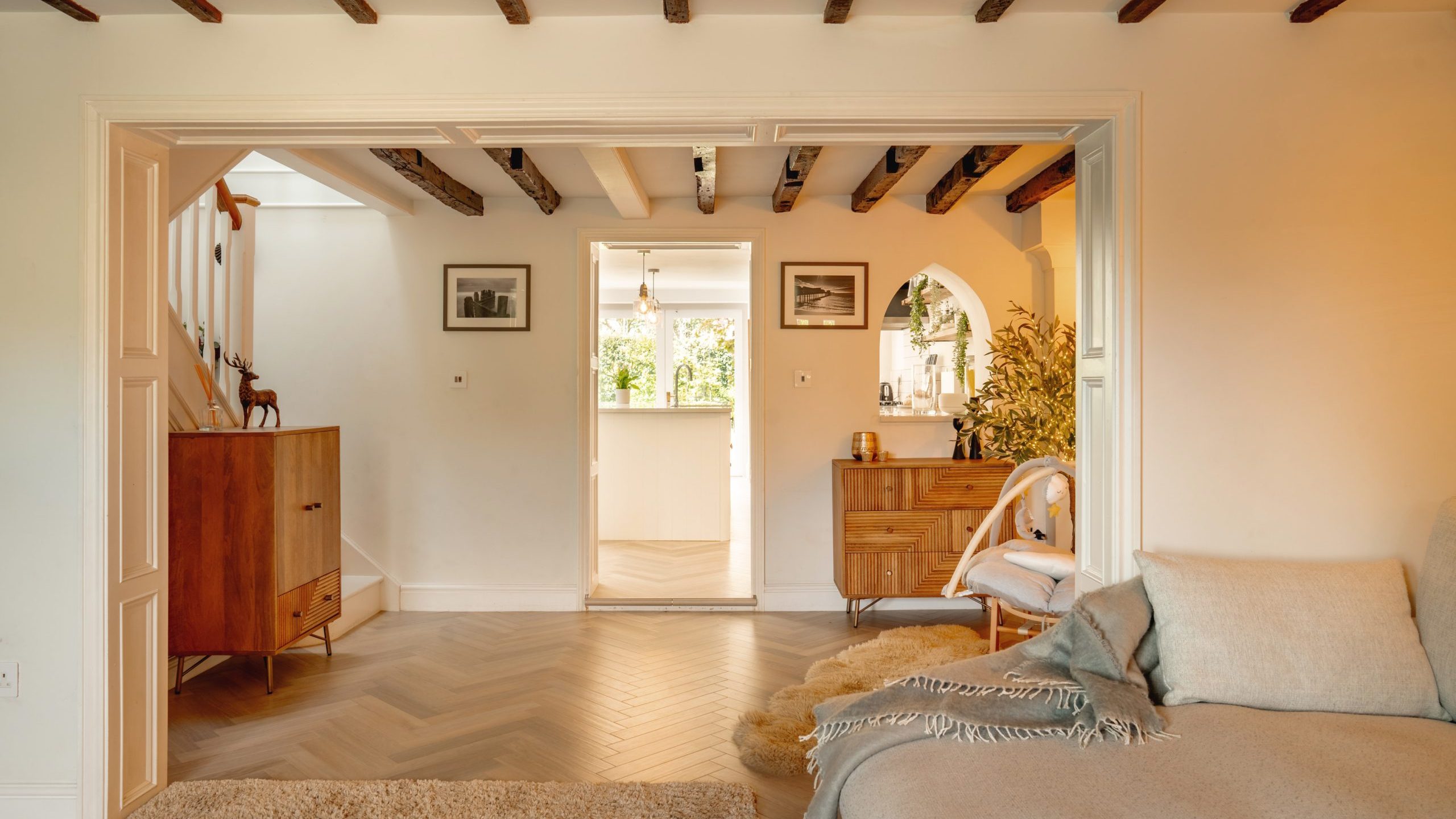 A cozy living room with a beige sofa, wooden furniture, and herringbone flooring. it features exposed wooden beams, leading through an open doorway to a bright kitchen.