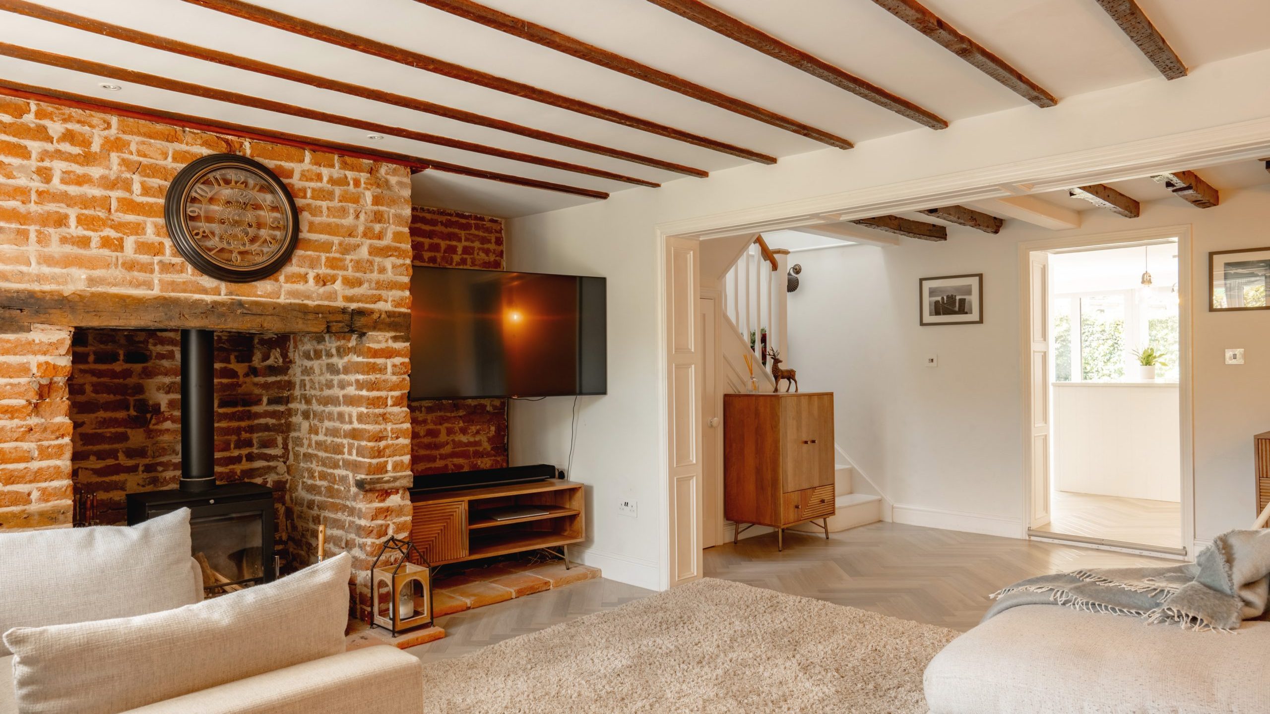 A cozy living room featuring exposed brick walls, wooden ceiling beams, and a fireplace. modern furniture and a mounted tv complement the rustic decor. two plush sofas and elegant flooring add to the ambiance.