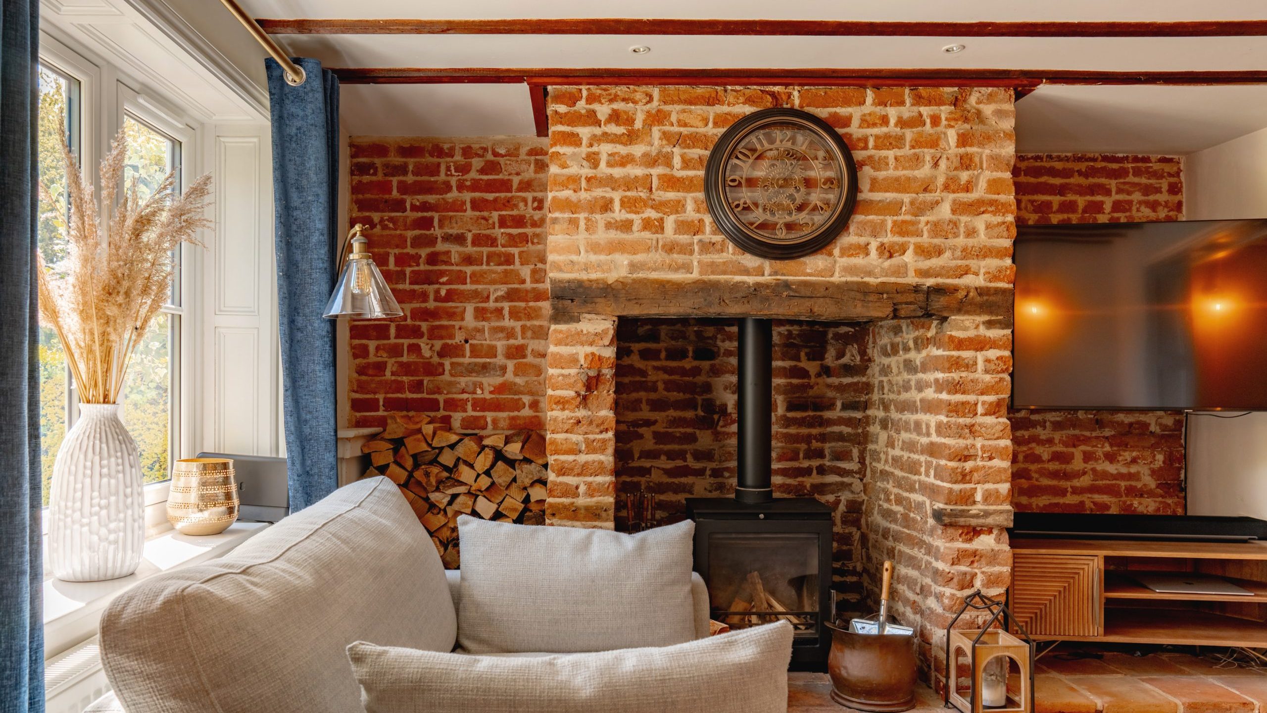 A cozy living room featuring exposed brick walls, wooden beams, and a wood-burning stove. there's a plush sofa, wooden shelves with a tv, and decor like a large clock and dried plants.