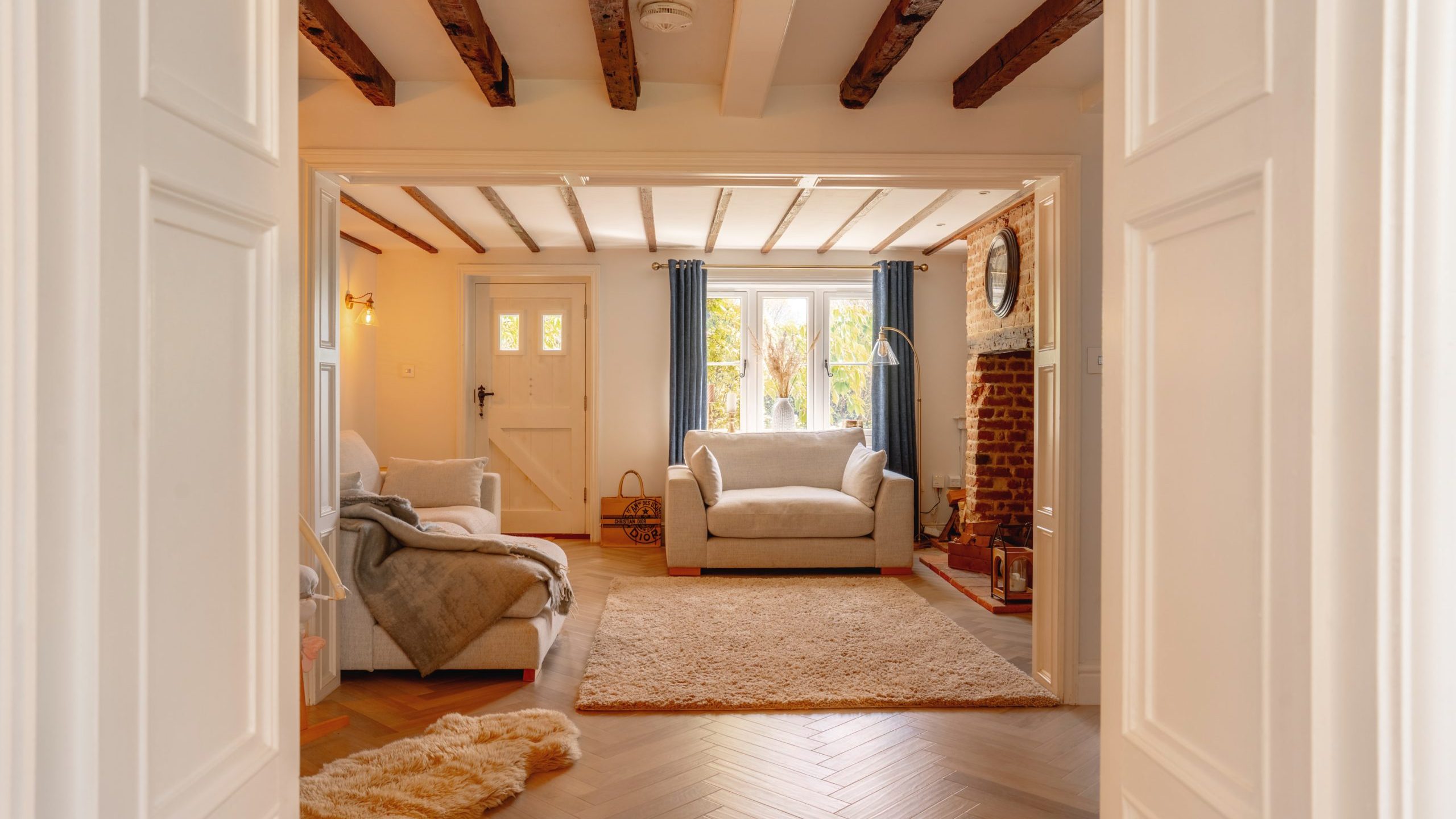A cozy living room viewed from an adjoining room, featuring exposed wooden beams, a white sofa, armchair, and a brick fireplace. a large window with curtains floods the space with natural light.