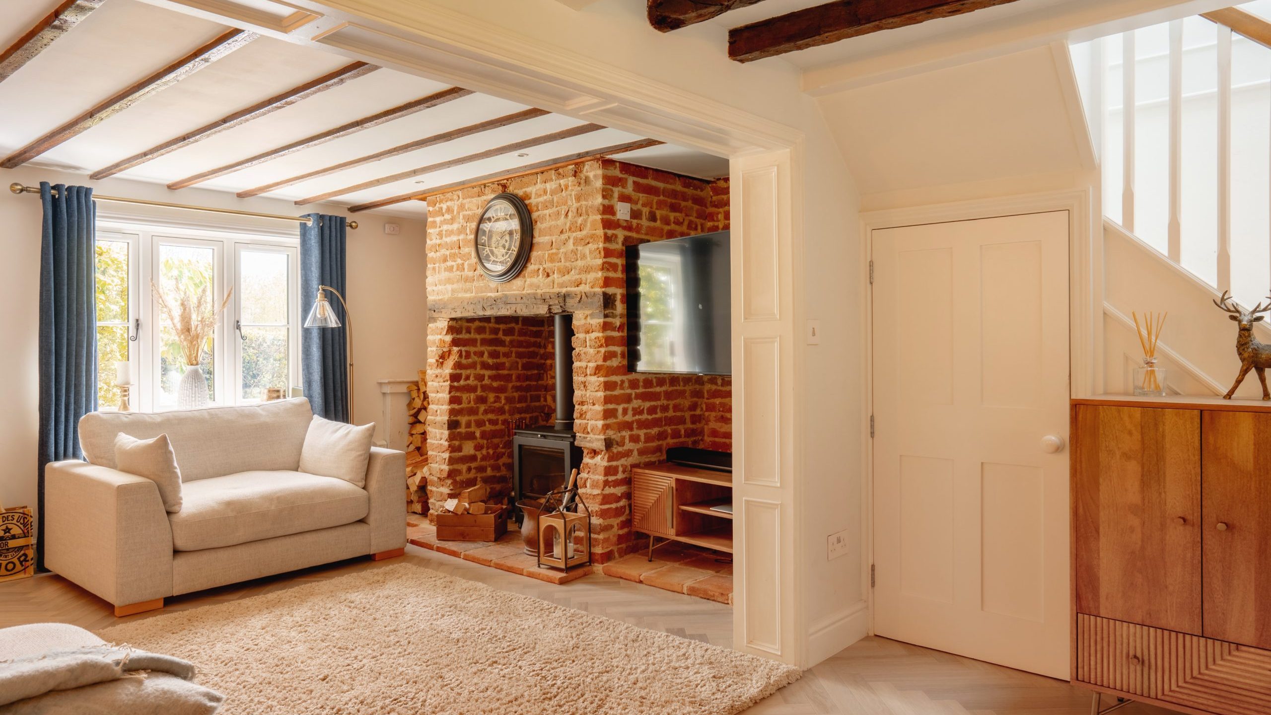 A cozy living room featuring exposed wooden beams, a brick fireplace with a mounted tv above it, a cream sofa, and large windows with curtains. there's also a plush rug and a wooden cabinet.