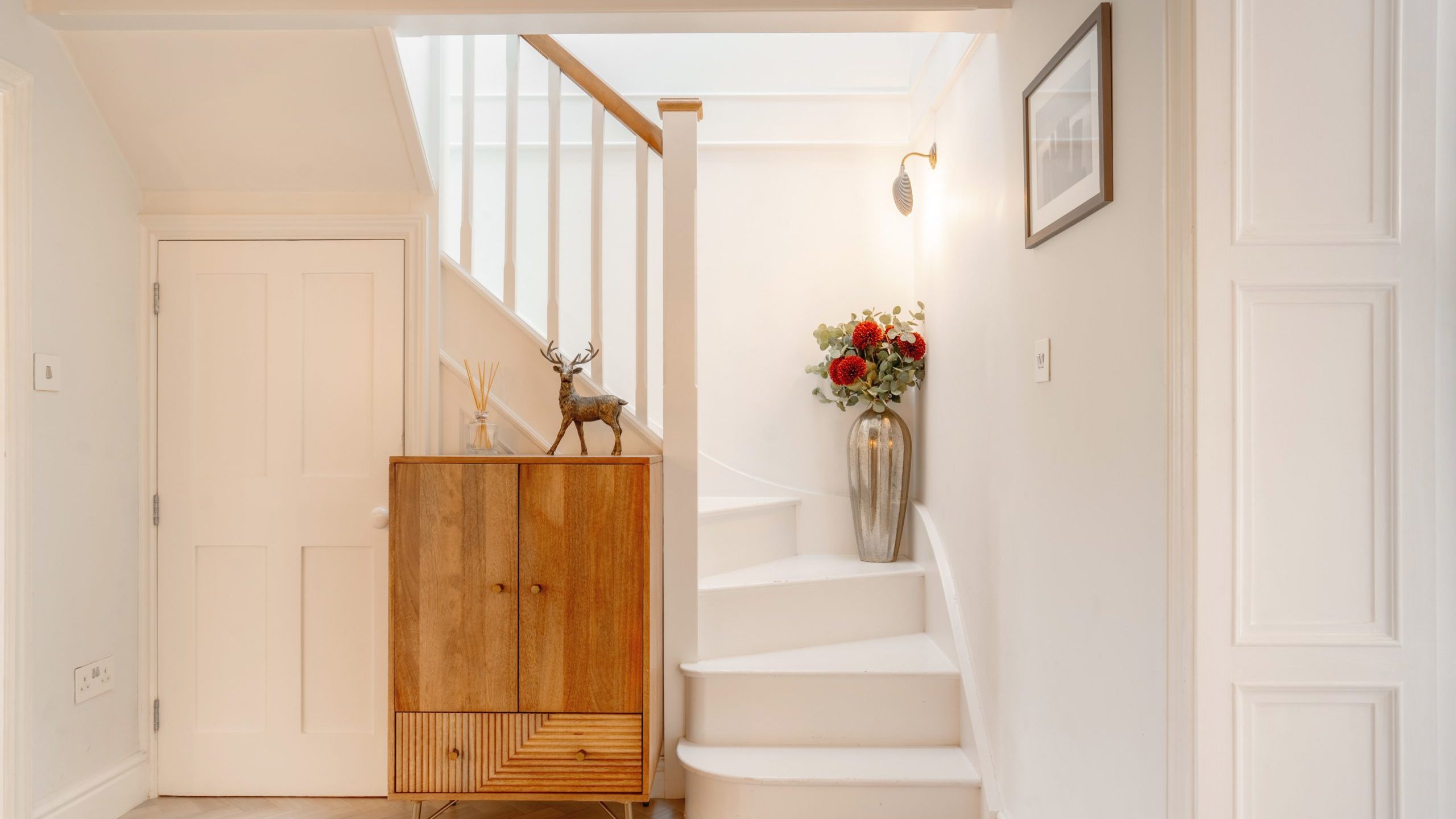 A cozy hallway featuring a wooden cabinet with a deer sculpture, vibrant flower bouquet and stairs leading up beside a wall with framed artwork. white doors and beige walls enhance the warm ambiance.