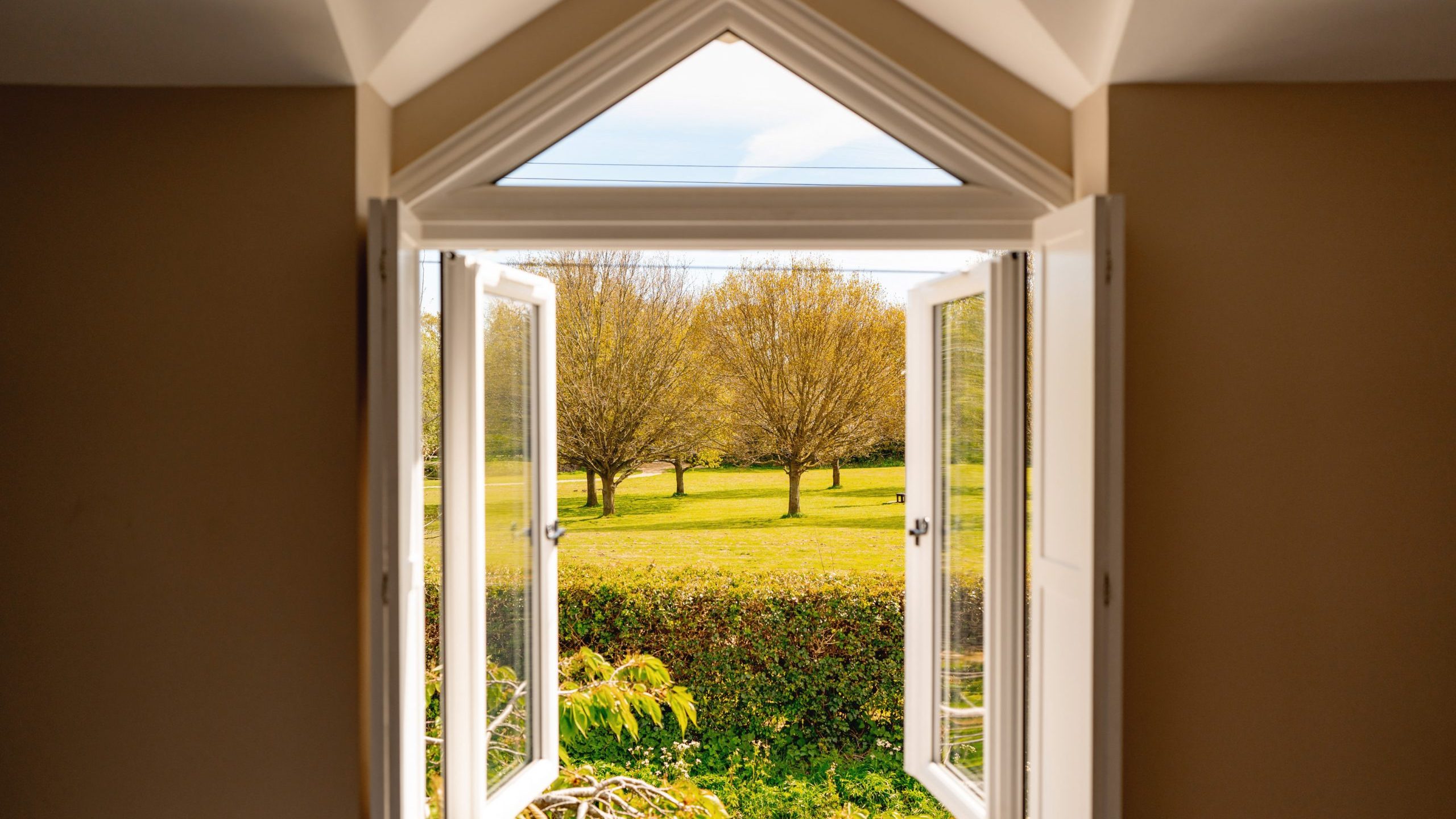 View from inside a room through an open triangular window, showing a lush green park with several trees and a bright blue sky.