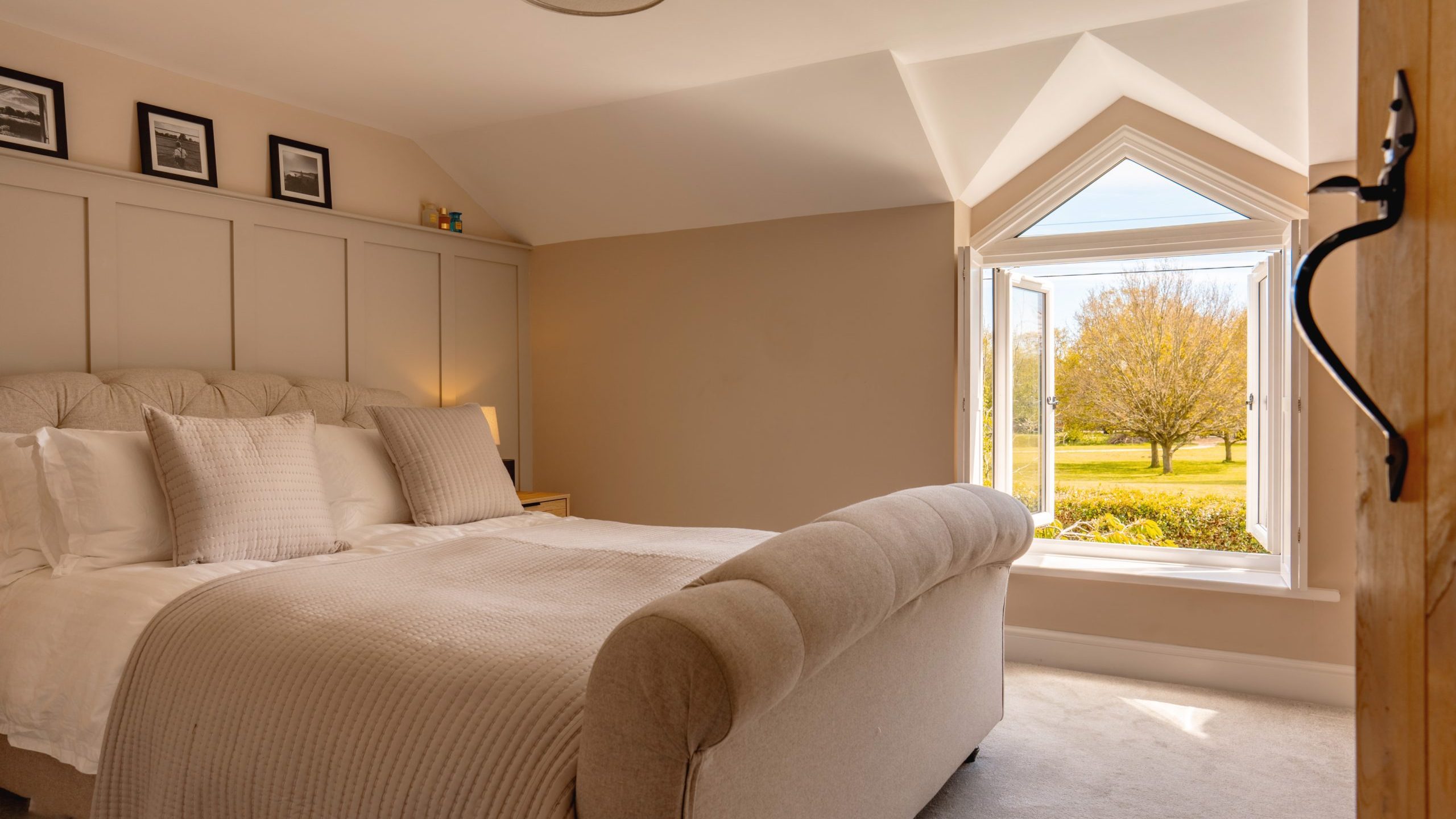 A well-lit spacious bedroom featuring a large bed with white bedding, a bench at the foot, several framed pictures on the wall, and a unique triangular window showing a view of a sunny landscape.