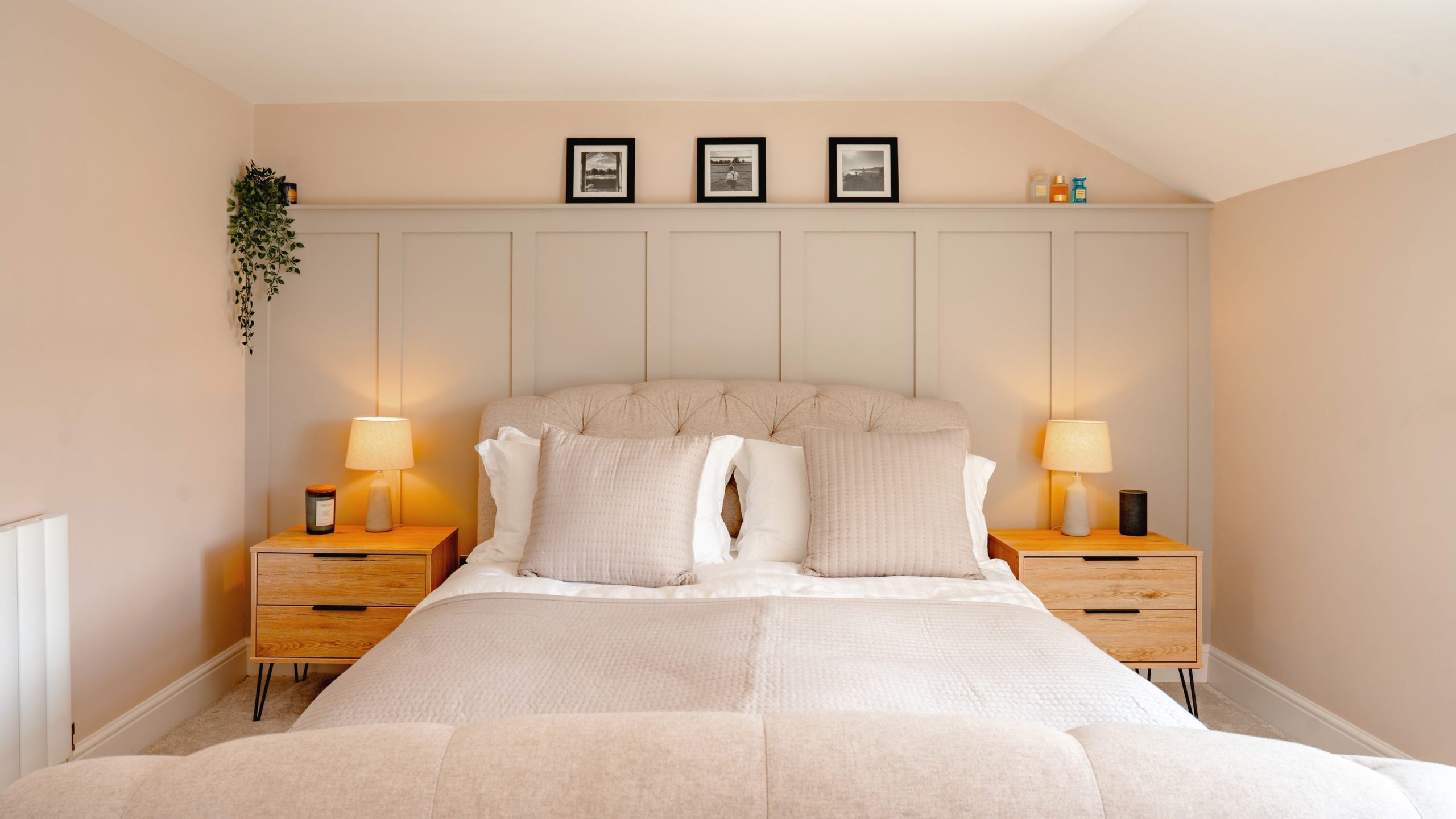 A tidy, well-lit bedroom featuring a large bed with a tufted headboard, flanked by two wooden nightstands with lamps. the room has soft beige walls and three framed pictures above the headboard.