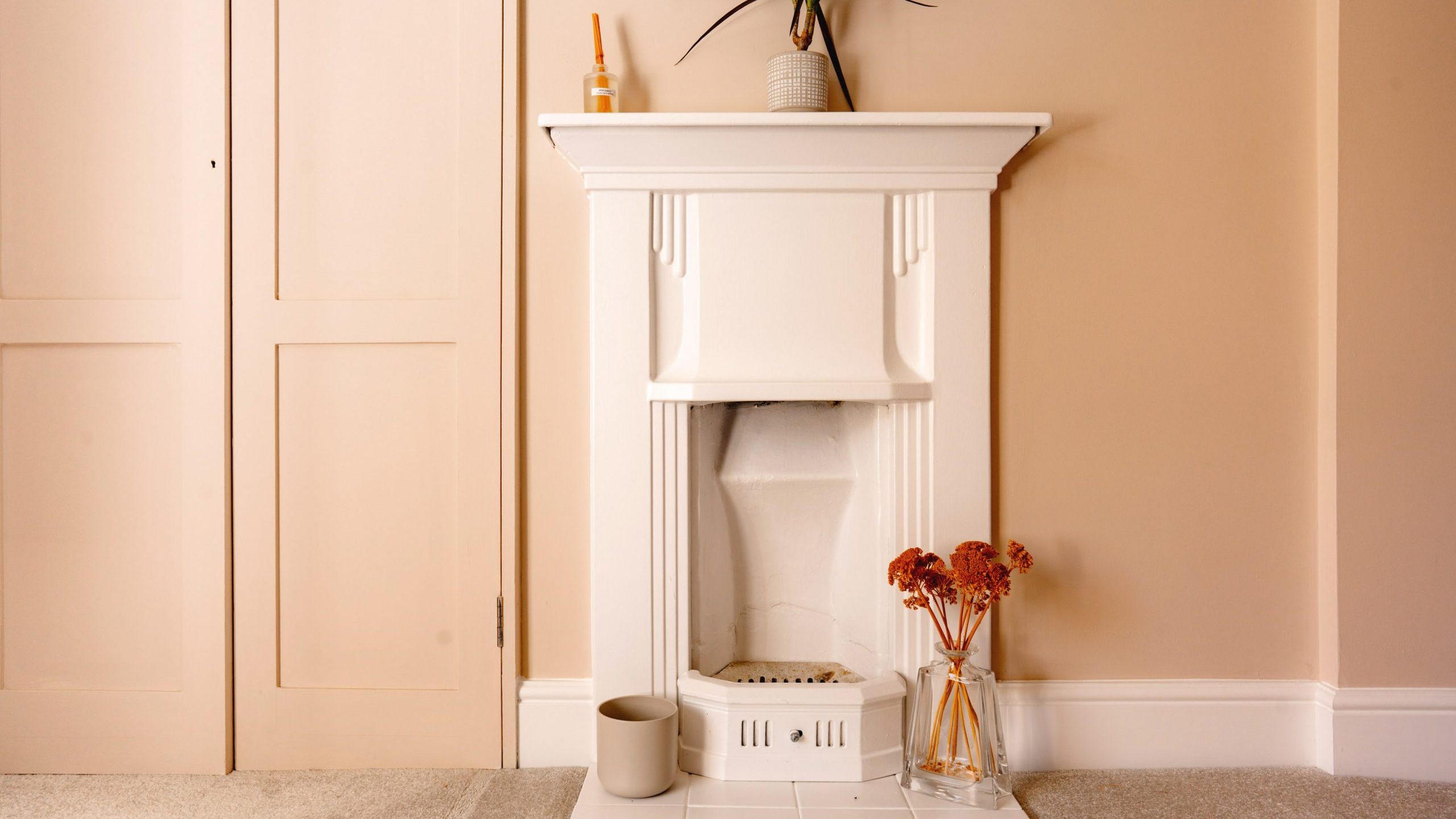 A cozy room corner featuring a decorative white fireplace with a modern vase and plant on top, flanked by pale orange flowers in a clear vase and essential oil diffusers on a neutral toned wall.