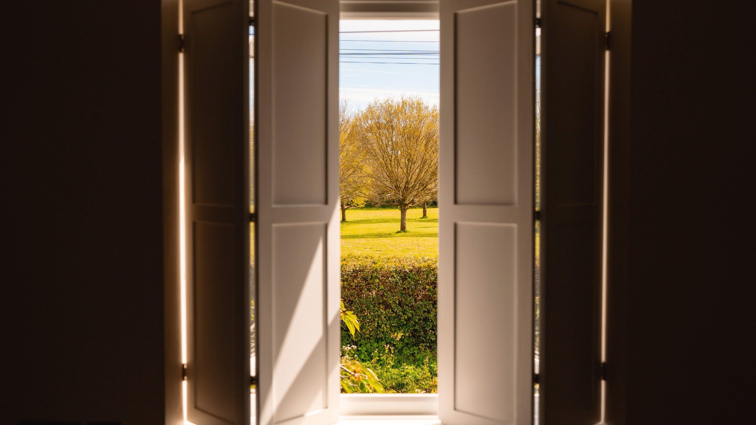 Open window with white shutters overlooking a sunny garden with lush green grass and scattered trees, framed by an interior view showing the window's deep sill and walls.