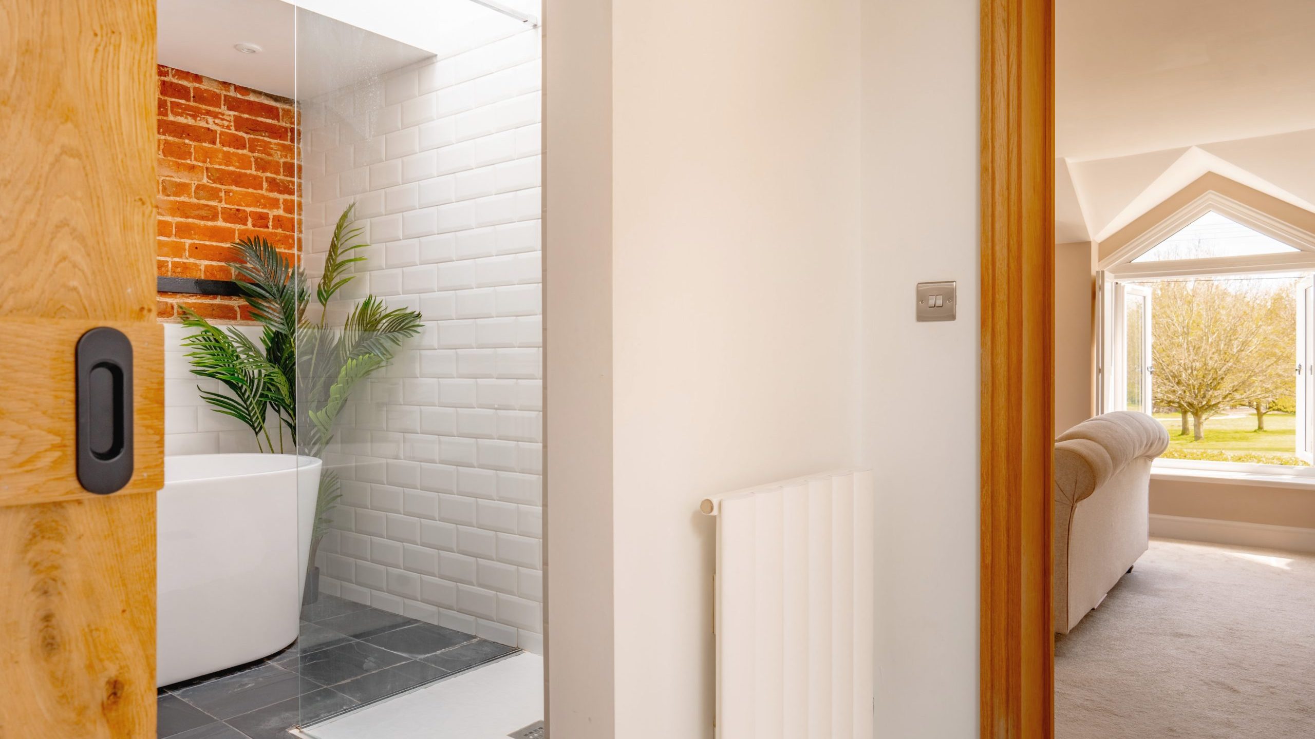 Modern bathroom interior viewed through a partially open wooden door, featuring white brick walls, a freestanding bathtub, and a tropical plant near a bright window.