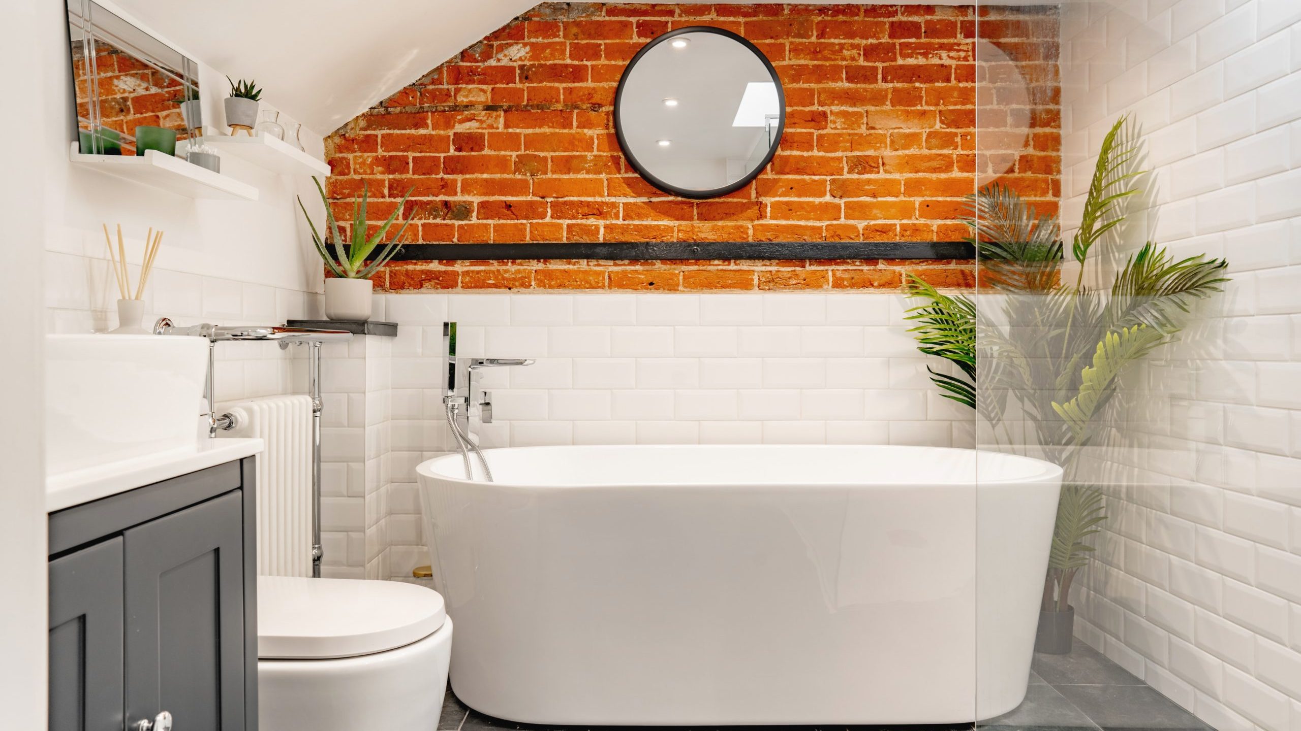 A modern bathroom featuring a freestanding white bathtub, exposed brick wall, and round mirror, with a toilet and gray vanity to the left, complemented by green plants and white subway tiles.