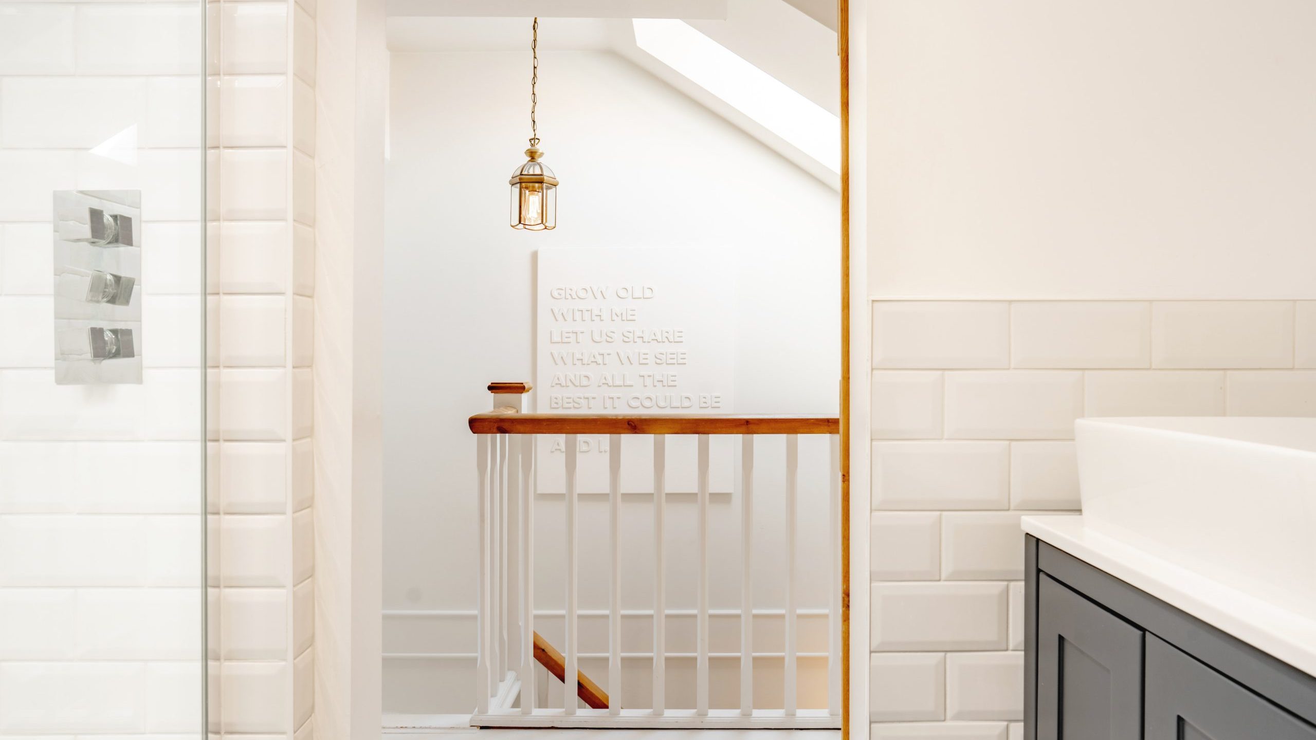 A bright, modern bathroom with white walls and tiles. a glass shower on the left and a dark gray cabinet to the right frame a small hallway leading to a wooden door. a stair railing is visible with text decorations on the wall.