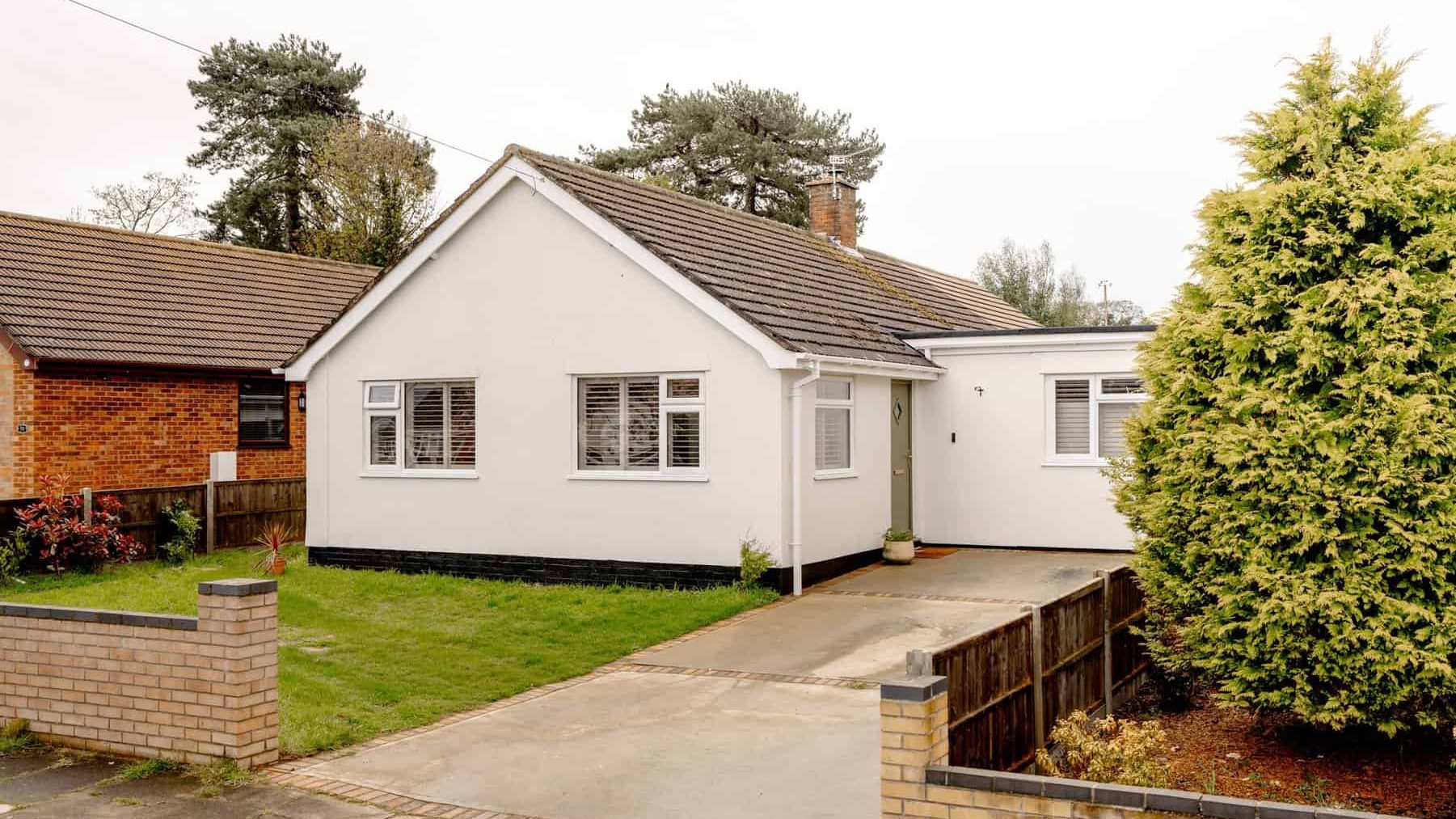 A white single-story house with black trim, featuring a driveway and surrounded by a brick fence, trees, and shrubs under a cloudy sky.