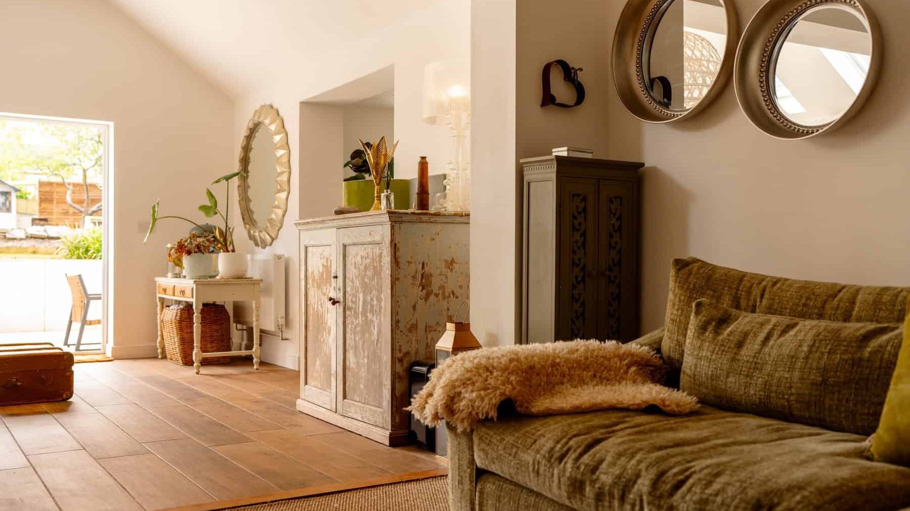 A cozy living room with a warm color palette, featuring a plush green sofa, rustic wooden furniture, and decorative mirrors on the wall. sunlight streams in, highlighting the serene and inviting space.
