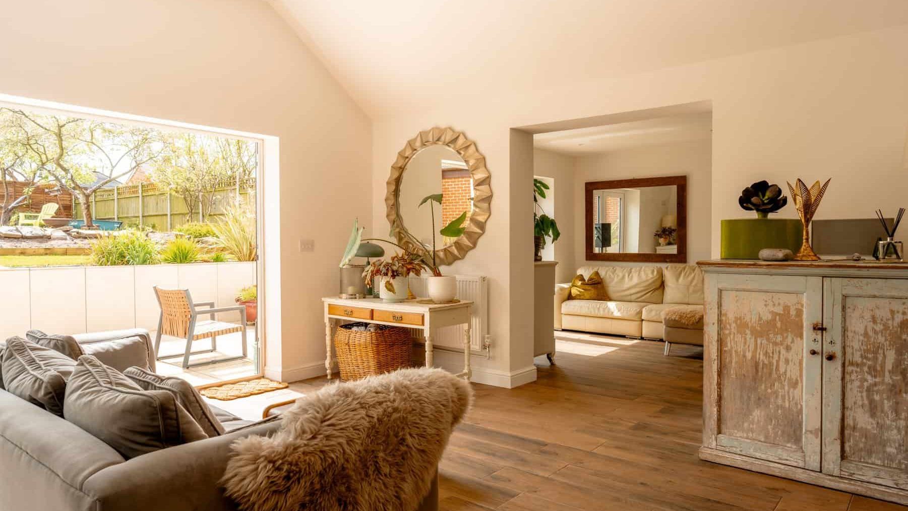 A cozy, well-lit living room with large windows showcasing a garden view. the room features a beige sofa, a rustic wooden cabinet, and decorative items. a round mirror and plants enhance the homely feel.