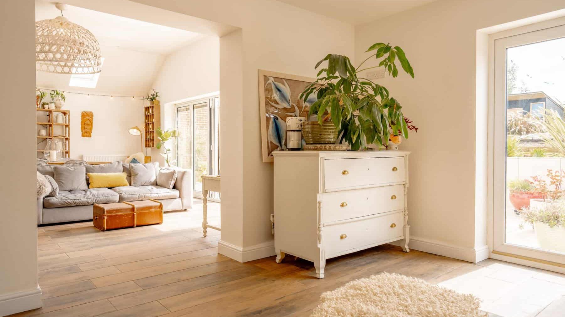 A bright and sunny living room with large windows, equipped with comfortable sofas and a chest of drawers adorned with plants and decorative items. a visible garden through the open door.