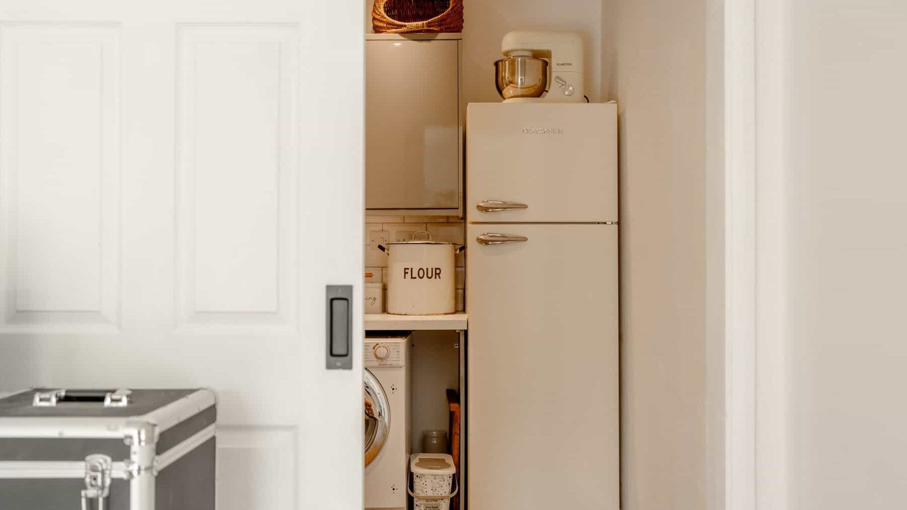 A small laundry room with a vintage white refrigerator, a washing machine, and shelving stocked with household supplies like flour and paint. the room is compact and neatly organized.