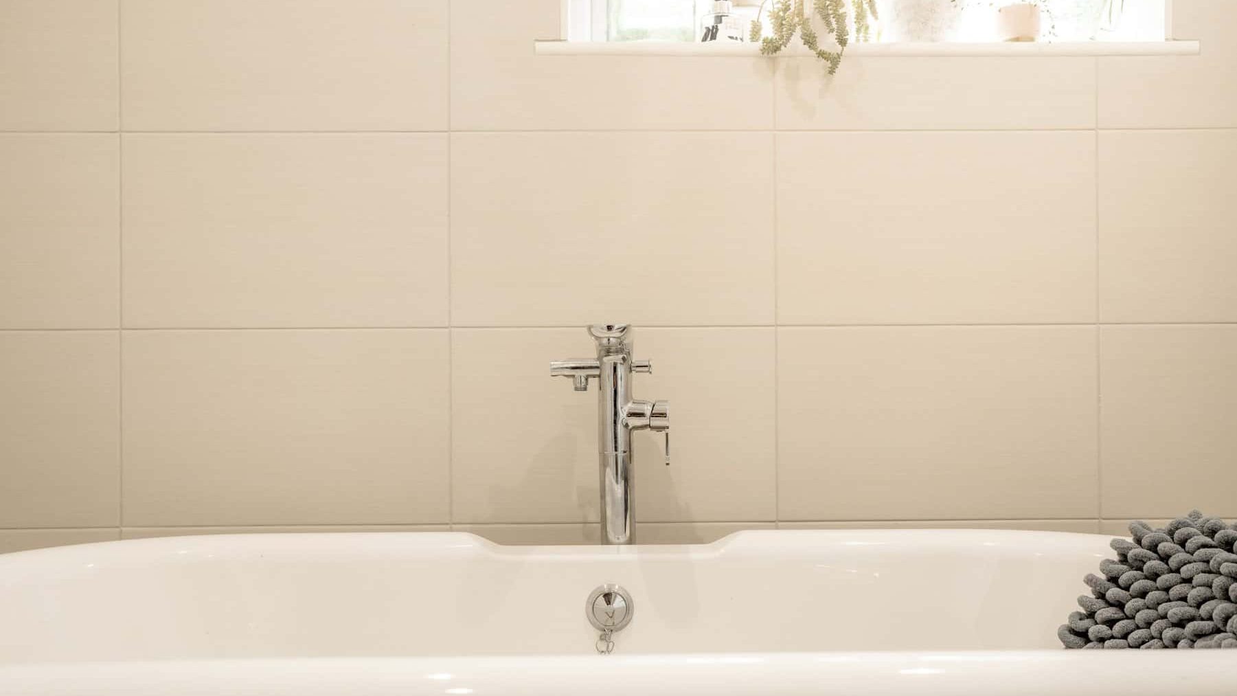 A modern, clean bathroom featuring a white bathtub with a sleek silver faucet. a window ledge above holds potted plants, enhancing a serene atmosphere. a stack of gray towels sits beside the tub.