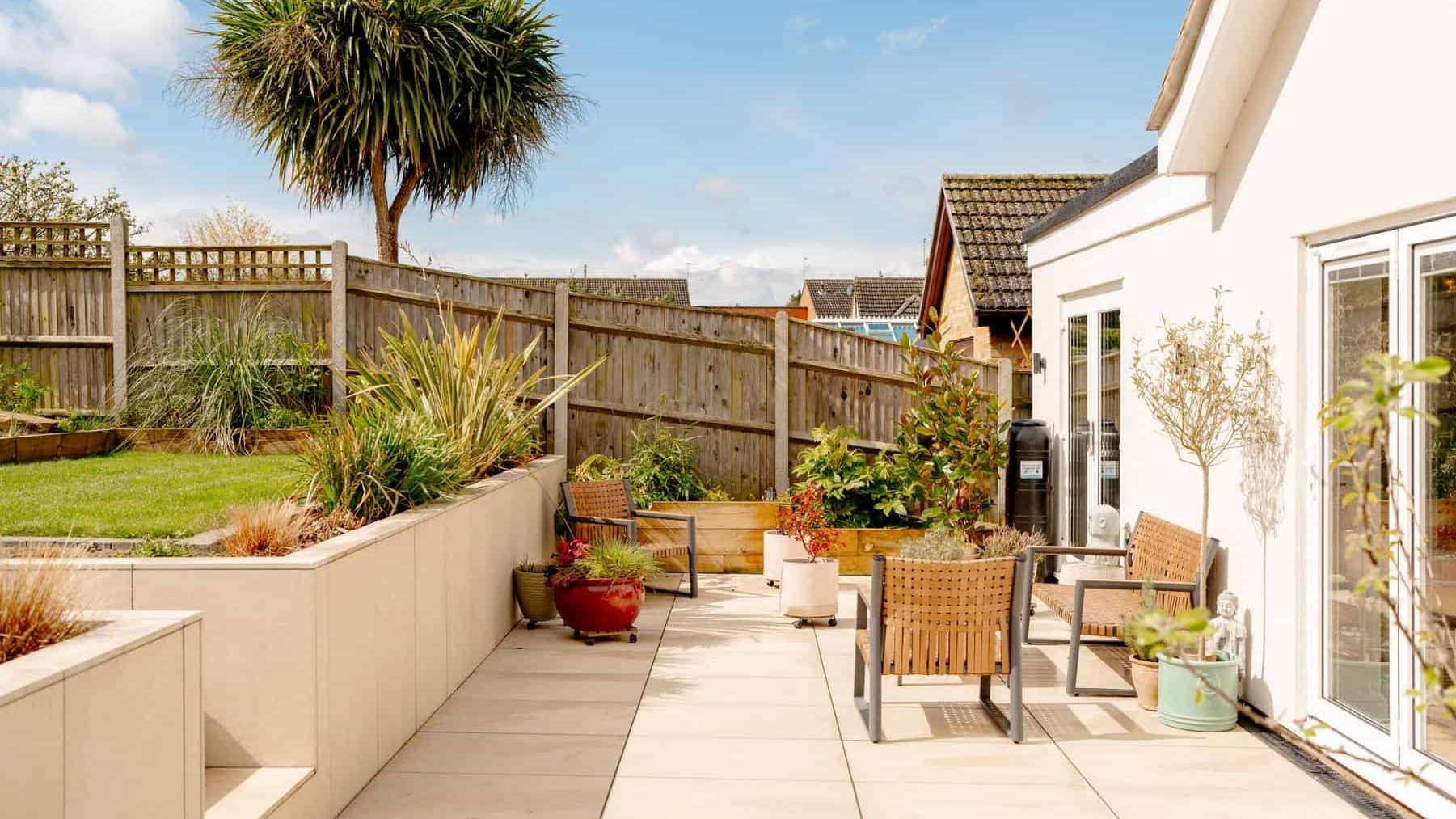A sunny backyard patio with tiled flooring, furnished with a wooden table and chairs. lush green plants and a small palm tree are visible, surrounded by a wooden fence.
